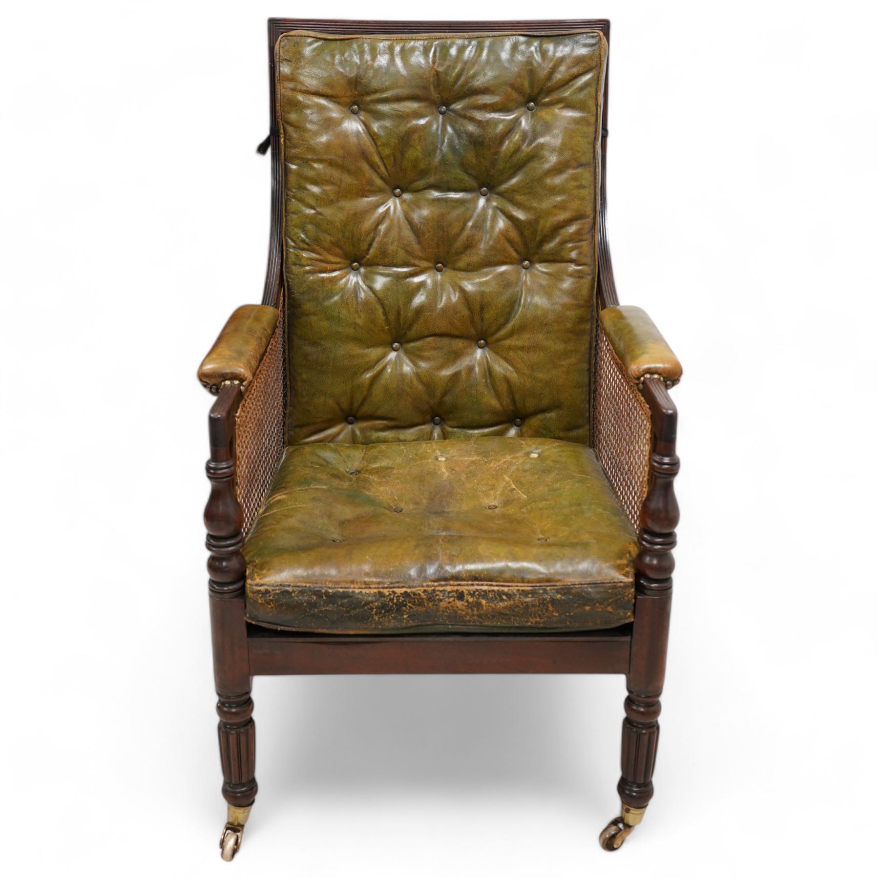 George IV mahogany framed library reading chair, reed moulded frame with cane work seat, back and sides, with loose cushions upholstered in green buttoned leather, upholstered arm rests, turned arm supports terminating to turned and reed carved feet, on castors 