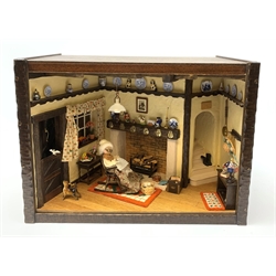 A 1/12th scale diorama of a cottage sitting room, depicting an elderly lady seated before a fireplace knitting, with dog and cat, in an interior furnished with blue and white plates, Staffordshire figures, toby jug, brass horse shoes, copper kettle, etc., H29cm L40cm D26cm.