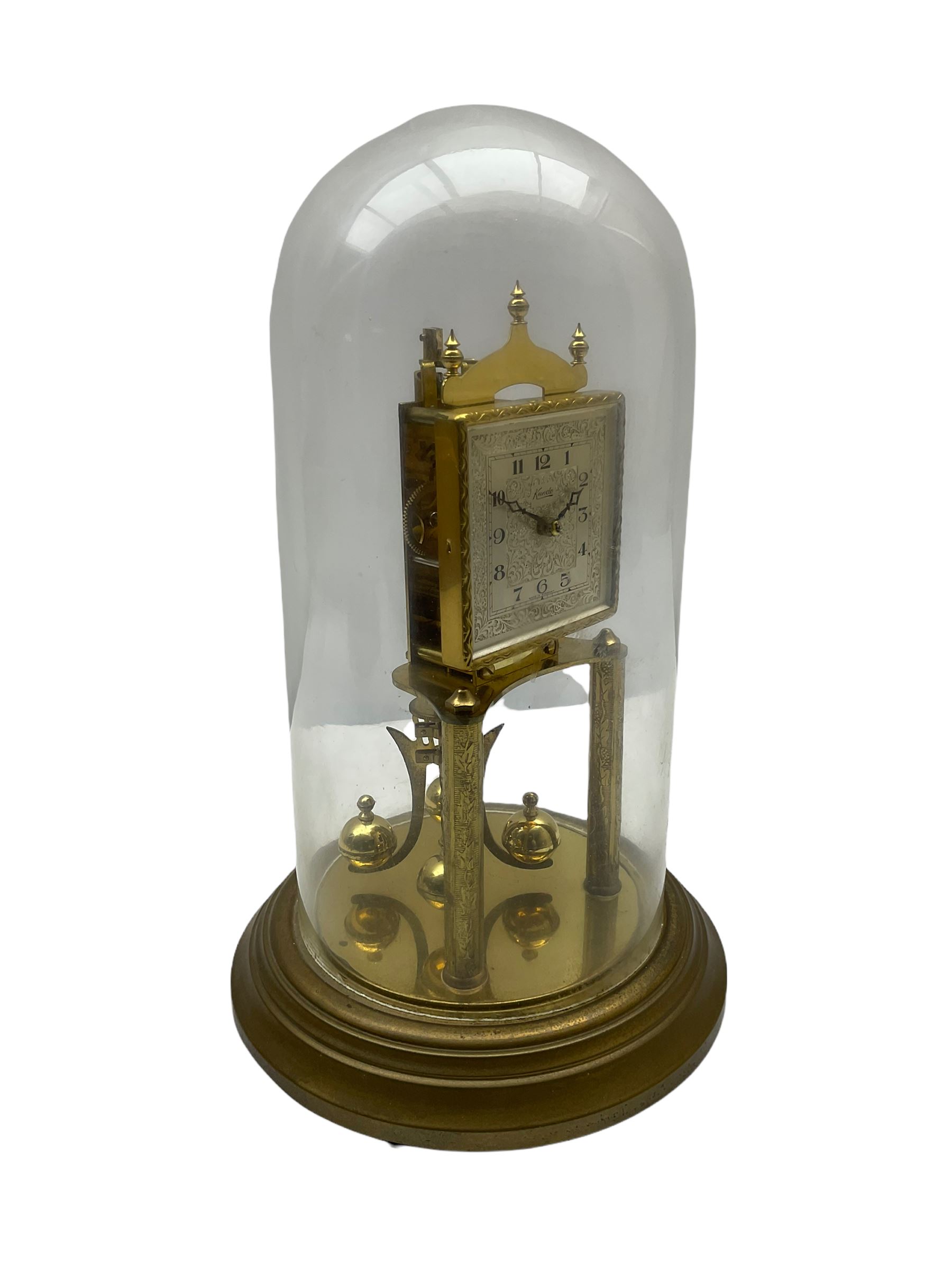 A “Kundo” 400-day torsion clock made by the 20th century German manufacturer Kieninger & Obergfell, with a  decorative square dial and pierced steel hands within a foliate bezel, on a circular base with adjustable feet, four-ball rotary pendulum under a glass shade. 
H30cm
