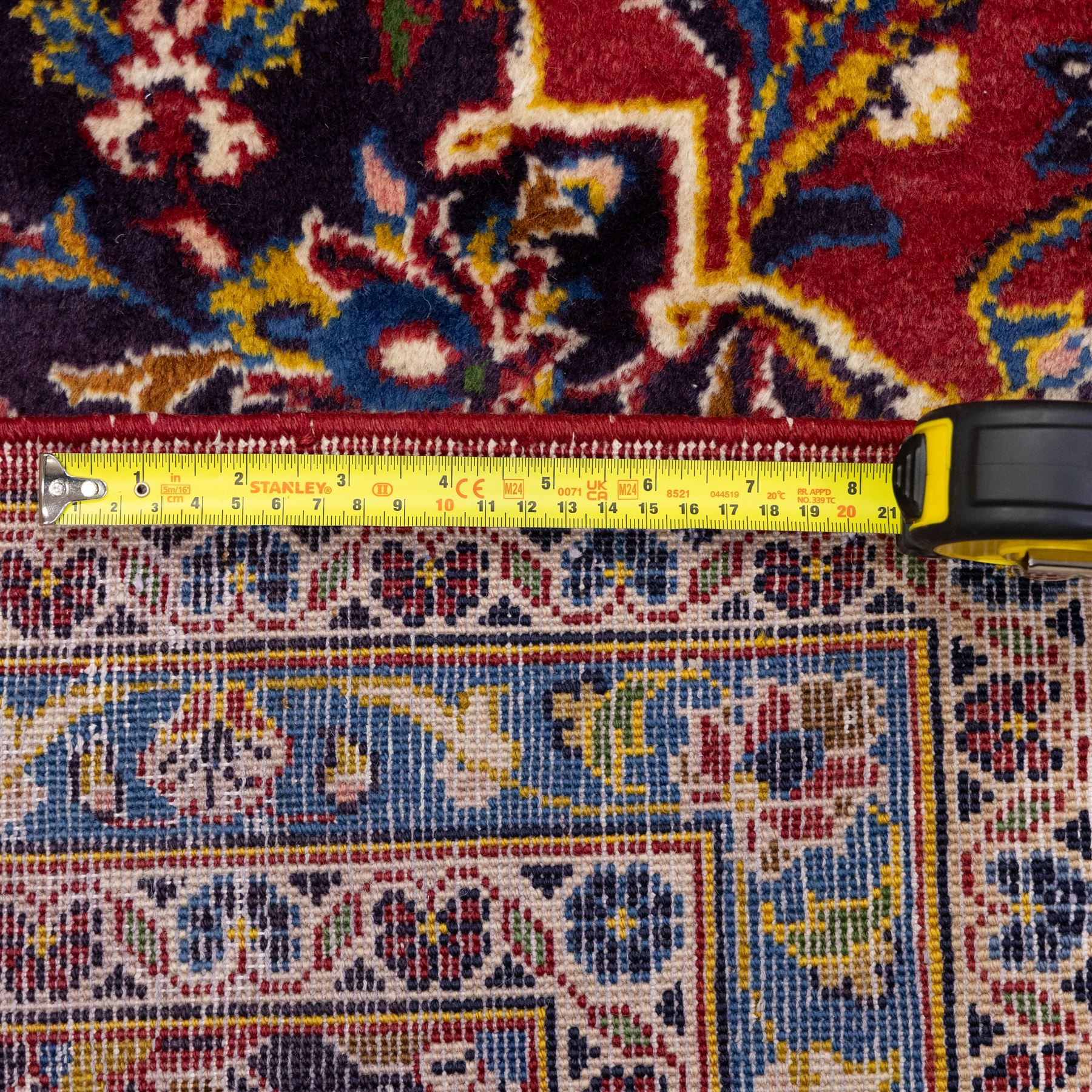 Central Persian Kashan hand-knotted carpet, crimson central field with lobbed medallion in navy blue and ivory ground, surrounded by a dense all-over floral design of scrolling vines, palmettes, and stylized blossoms, enclosed within a deep navy main border, flanked by multiple guard stripes with fringe ends 


