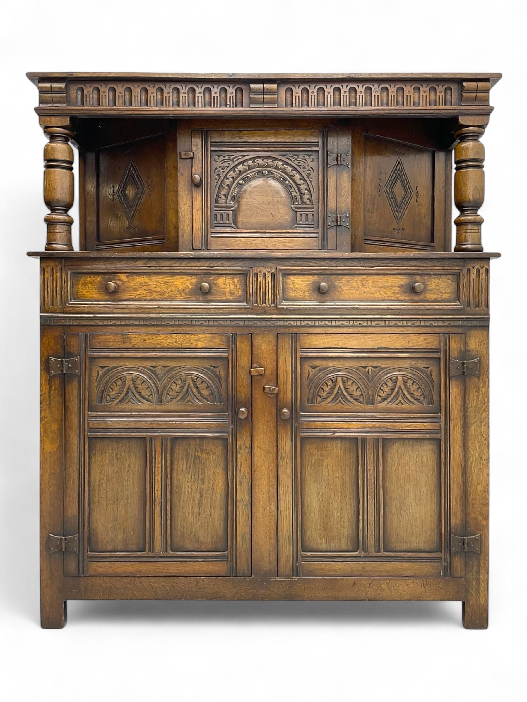 Jacobean design oak court cupboard, upper section with carved arched panelled cupboard door flanked by turned columns, beneath carved and moulded frieze, lower section fitted with two drawers over two cupboard doors with gothic arched panel decoration, raised on stile supports
