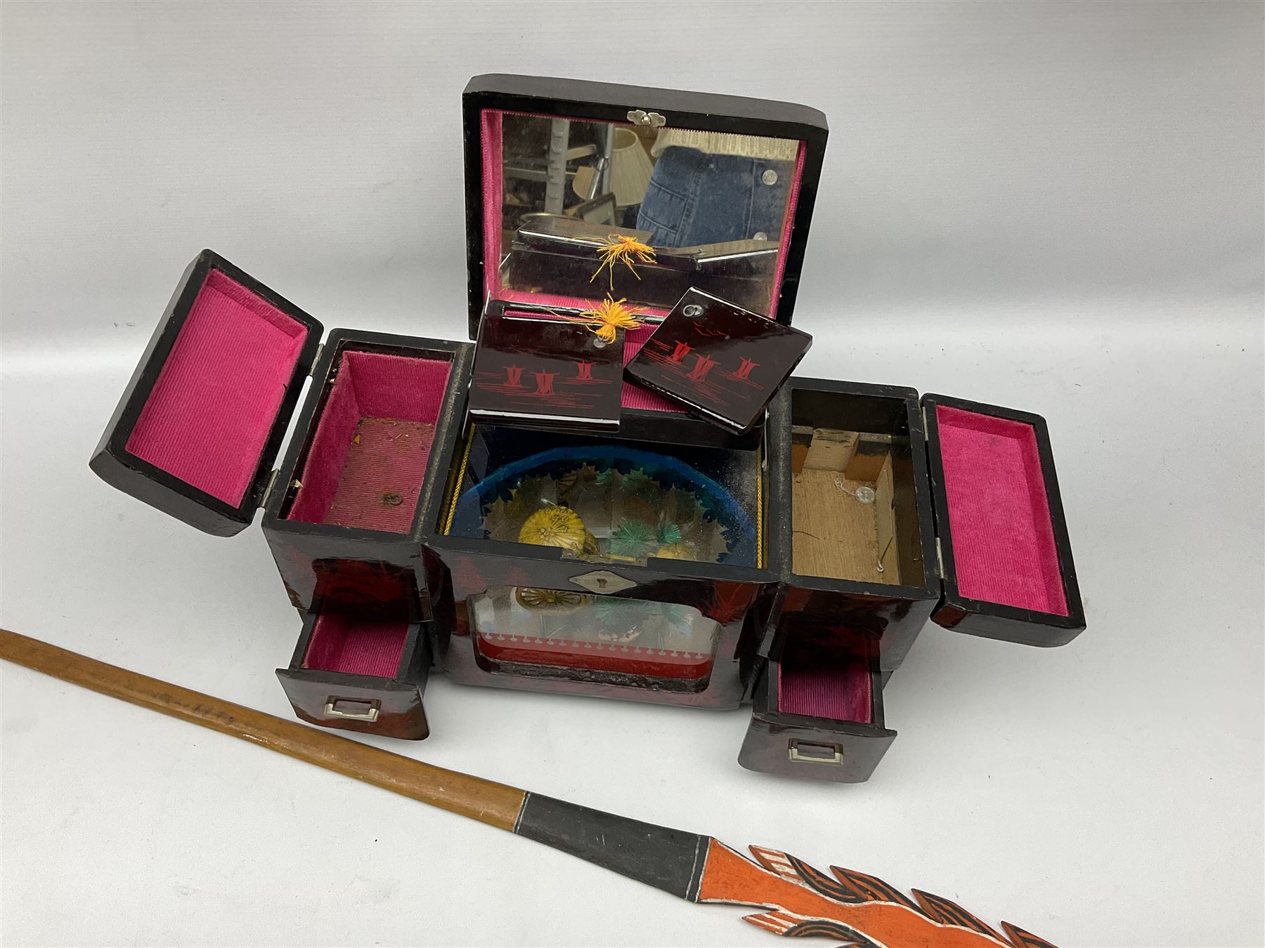 Oriental lacquered jewellery box, the body decorated with a black and red mountainous riverscape enclosing a glass compartment with simulated ivory rickshaw figure and three hinged lids opening to reveal mirror and various compartments and painted ceremonial stick