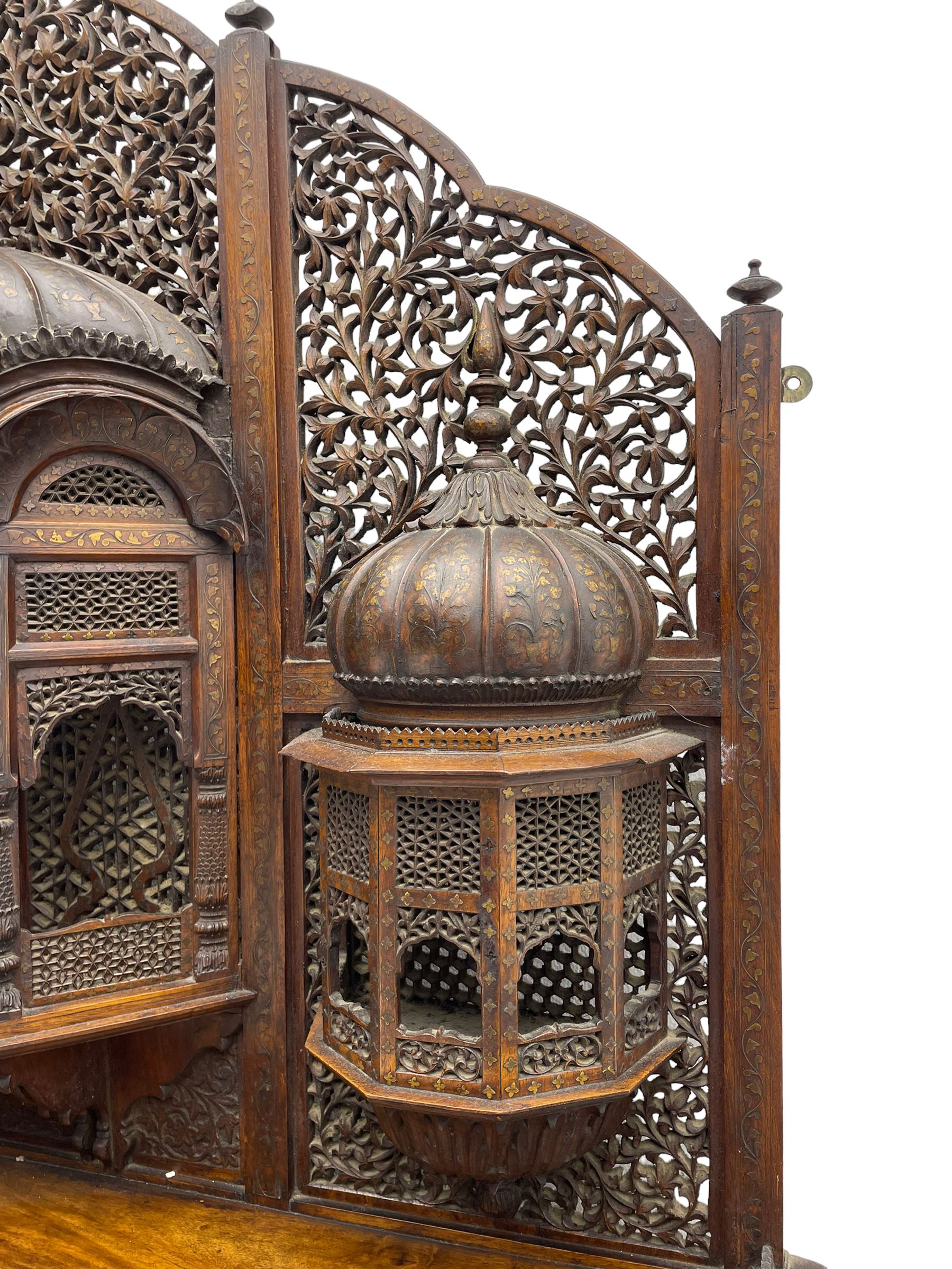 Late 19th century Indian Burmese carved and pierced hardwood and brass inlaid wall shelf, the shaped back profusely decorated with trailing and interlaced foliage, with projecting architectural structures, the central balcony with canopy top and five open windows with arched apertures on columns, two flanking five-sided balconies, the shelf supported by six foliate carved columns with pierced cusped pointed arches 