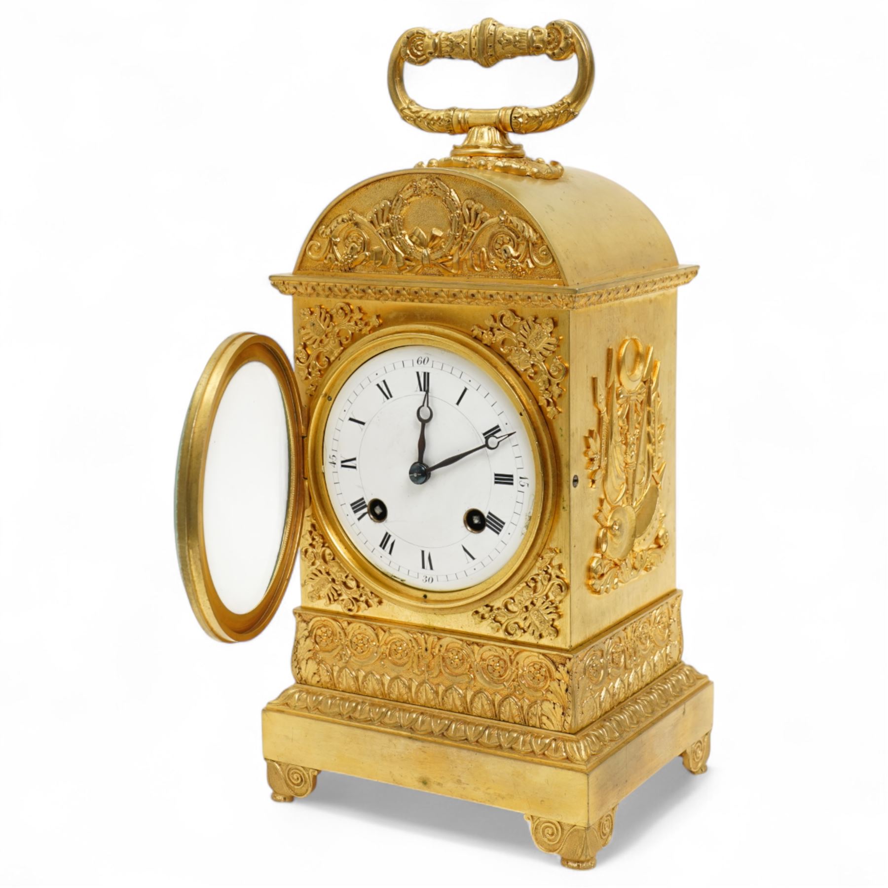 Fine Swiss ormolu 8-day early 19th century Pendule d'Officer style clock c1820,  circular double train ting-tang movement striking the quarters on a graduated pair of bells and sounding the hour on one, going train with a vertical platform lever escapement and over sprung balance, rack striking, with visible hour and quarter strike work to the backplate, movement unsigned, convex enamel dial with Roman numerals and steel moon hands behind a glazed gilt bezel, case with a scroll cast handle to the arched top with applied rococo decoration to the tympanum, both sides of the case adorned with a military trophy of arms motif and deep frieze with foliate arcading beneath, on a rectangular plinth with decorative scroll engraved bracket feet. With Key.   