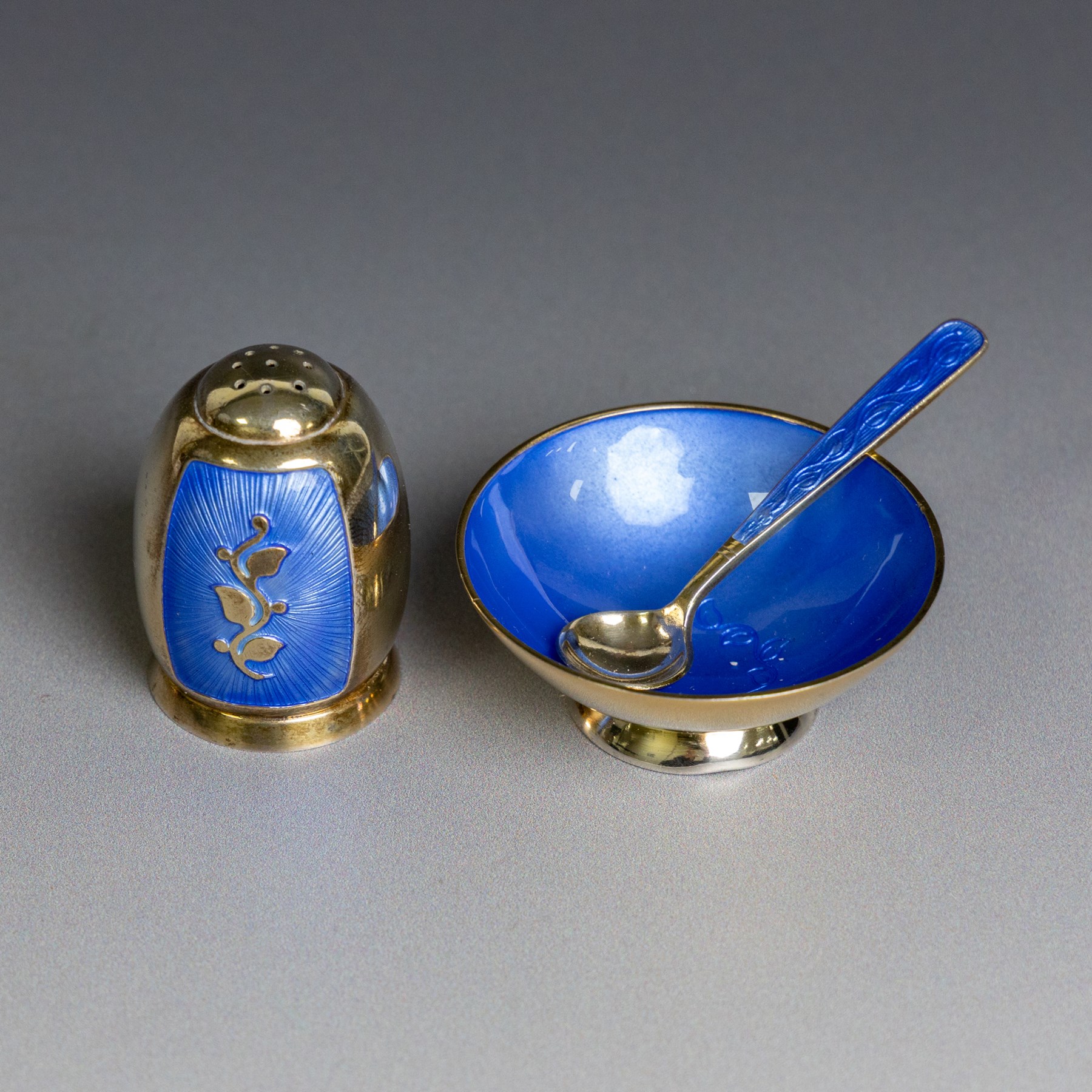 Mid 20th century Danish silver-gilt and guilloche enamel cruet set by Volmer Bahner, comprising pepper shaker, open salt and salt spoon, the spoon handle and bowl interior with blue basse taille enamel and vine motif, the pepperette with panels of blue guilloche enamel with similar applied vine decoration, all stamped Sterling Denmark VB, pepperette H4cm
