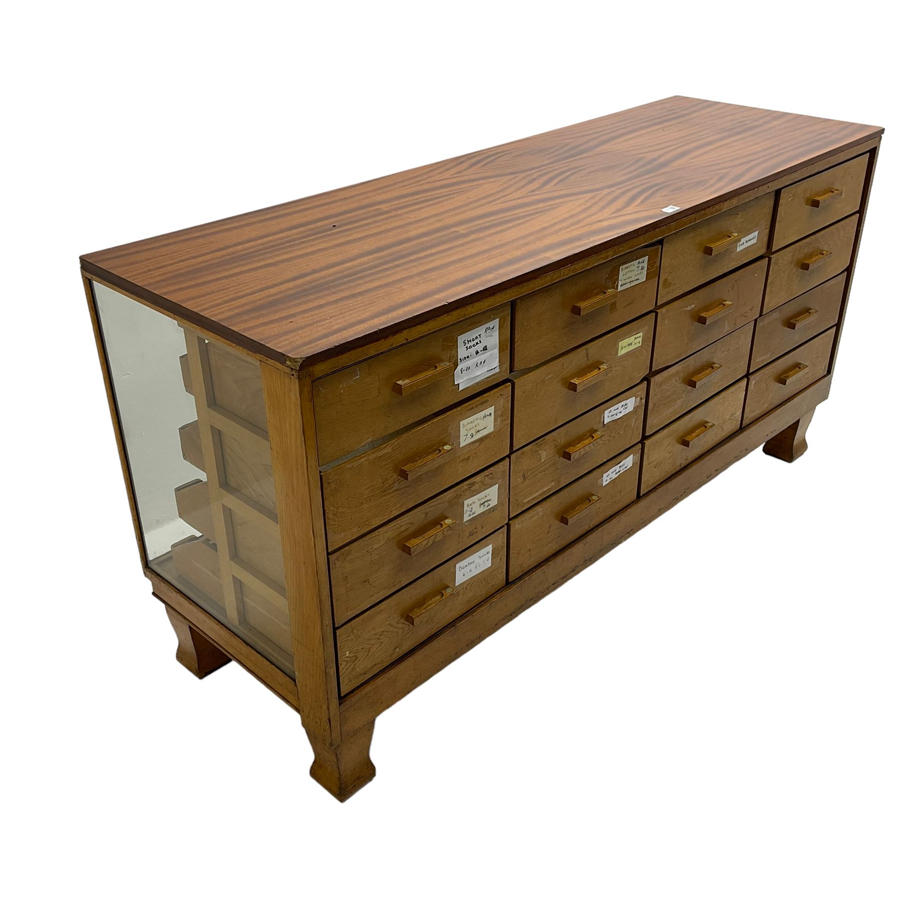Early 20th century oak framed haberdashery shop counter, veneered top with glazed front and sides, fitted with sixteen graduating drawers with oak fronts and handles, raised on shaped supports