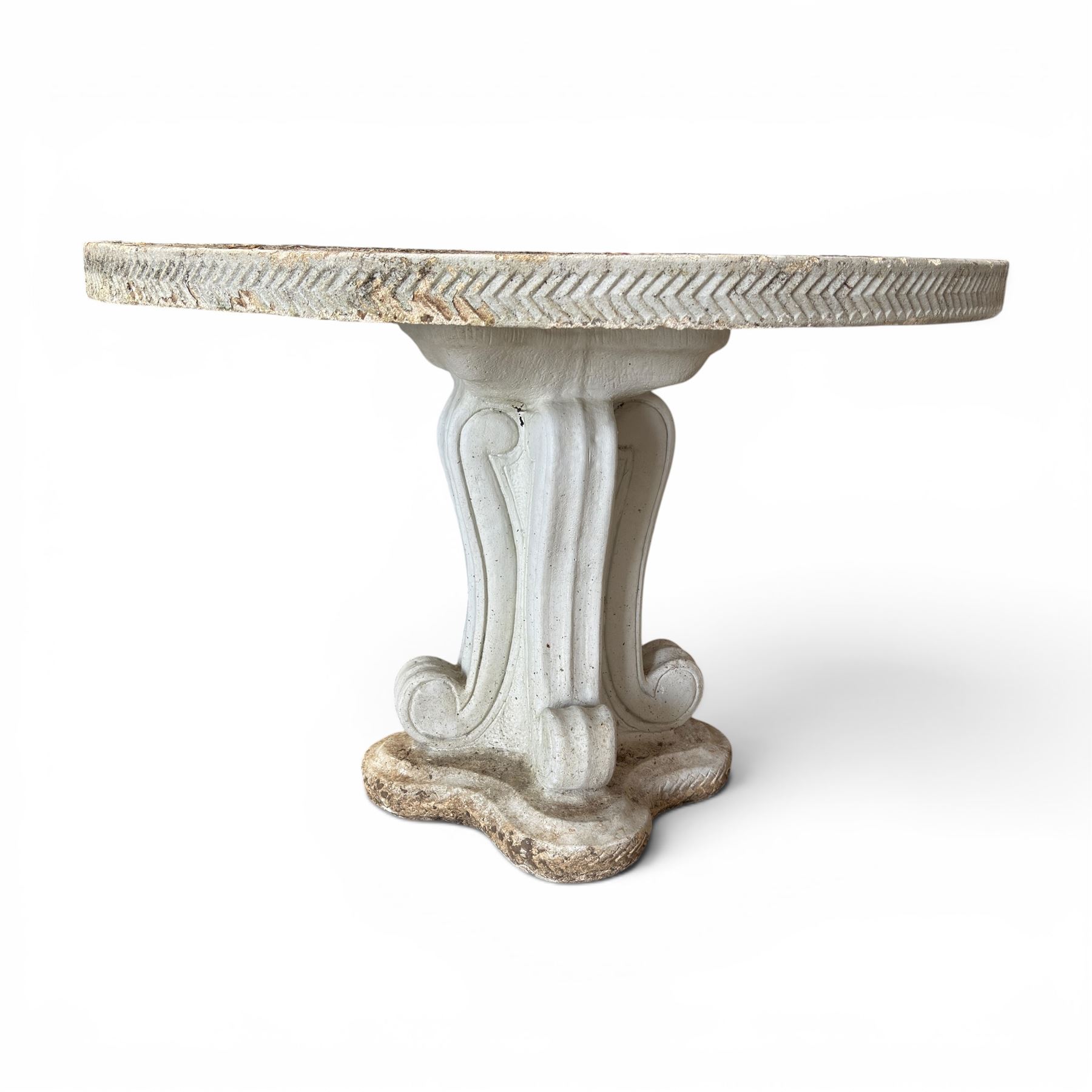 Weathered cast stone garden table, circular top etched with central floral medallion, the edge with repeating chevron motifs, raised on a quadriform S-scroll pedestal with stepped and chevron carved base
