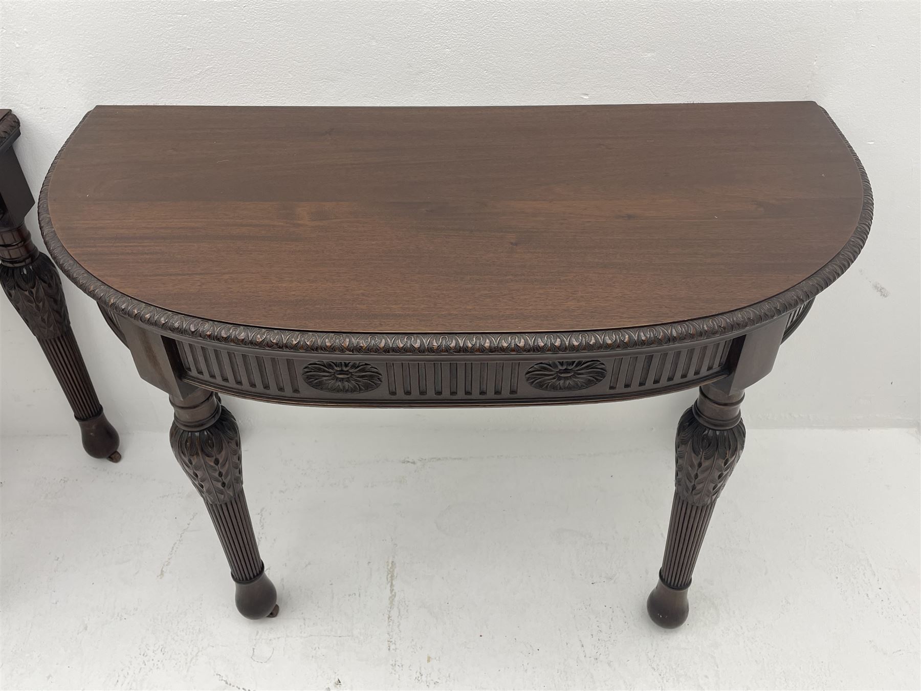 Pair early 20th century Hepplewhite style mahogany D-end console tables, the curved top with foliate carved edge, fluted frieze rails carved with flower heads, turned and reeded acanthus supports with recessed castors