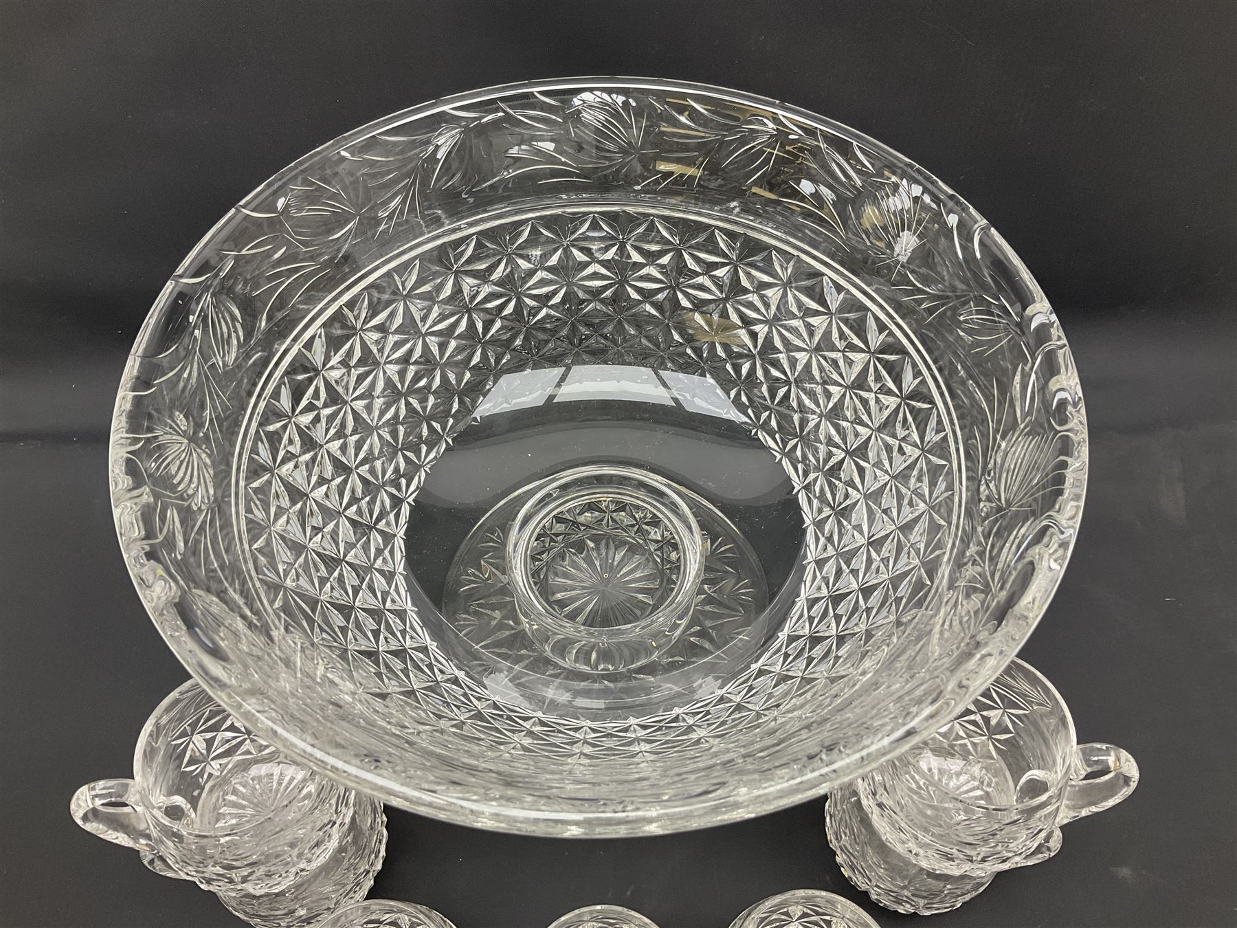 Large Stuart crystal pedestal punch bowl, of circular form, the rim with engraved floral decoration, upon spreading circular foot with radial cut decoration, with seven matching Stuart crystal drinking glasses, each with a C handle, each acid signed beneath, bowl H20cm