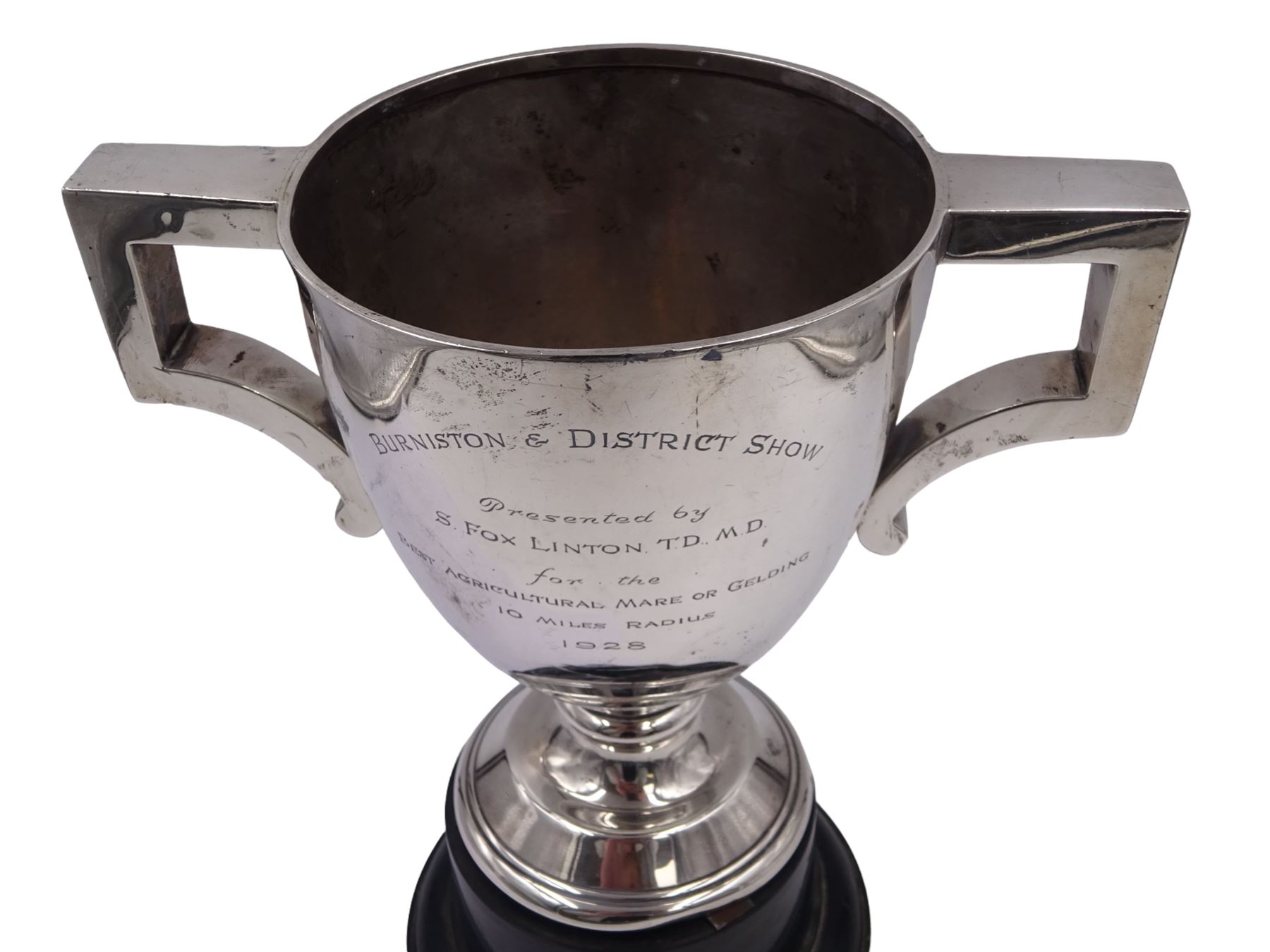 1920s silver trophy cup, with angular twin handles, upon knopped stem with circular spreading foot, the body with presentation engraving 'Burniston & District Show, presented by S Fox Linton TD MD, for the Best Agricultural Mare or Gelding, 10 miles radius, 1928' and engraved with winners verso, hallmarked Charles Boyton & Son 1921, upon black plastic base, including base 25cm
