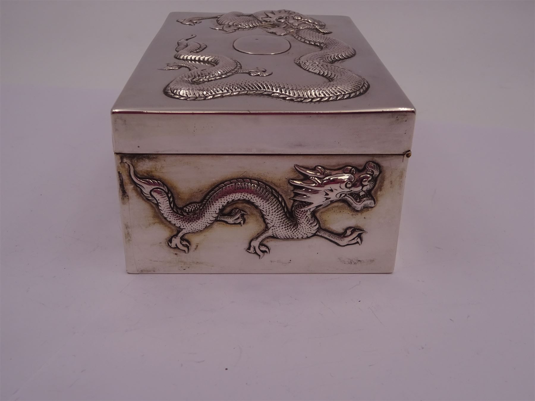 Late 19th century/early 20th century Chinese export silver mounted cigarette box, of rectangular form, decorated in relief with dragons, the hinged cover with similar dragon decoration and central blank oval cartouche, opening to reveal a softwood lined compartmentalised interior, stamped beneath with character mark and KMS for maker Kwong Man Shing, H6.3cm, W15.5cm, D9.5cm