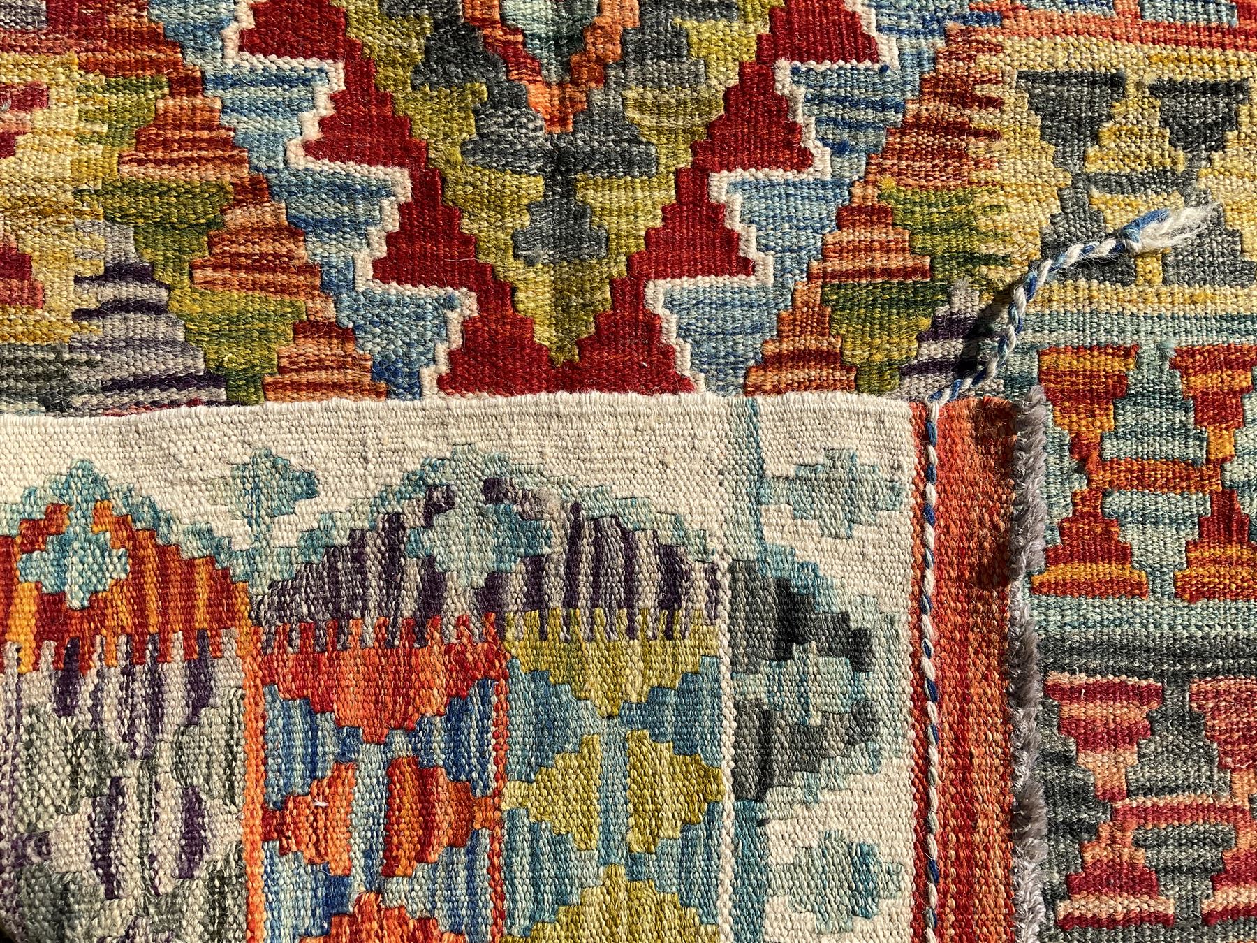 Afghan Chobi Kilim ivory ground rug, central geometric design consisting of stacked lozenge medallions in shades of red, green, and blue, flanked by multicoloured triangular motifs, guarded by zigzag patterns in contrasting tones