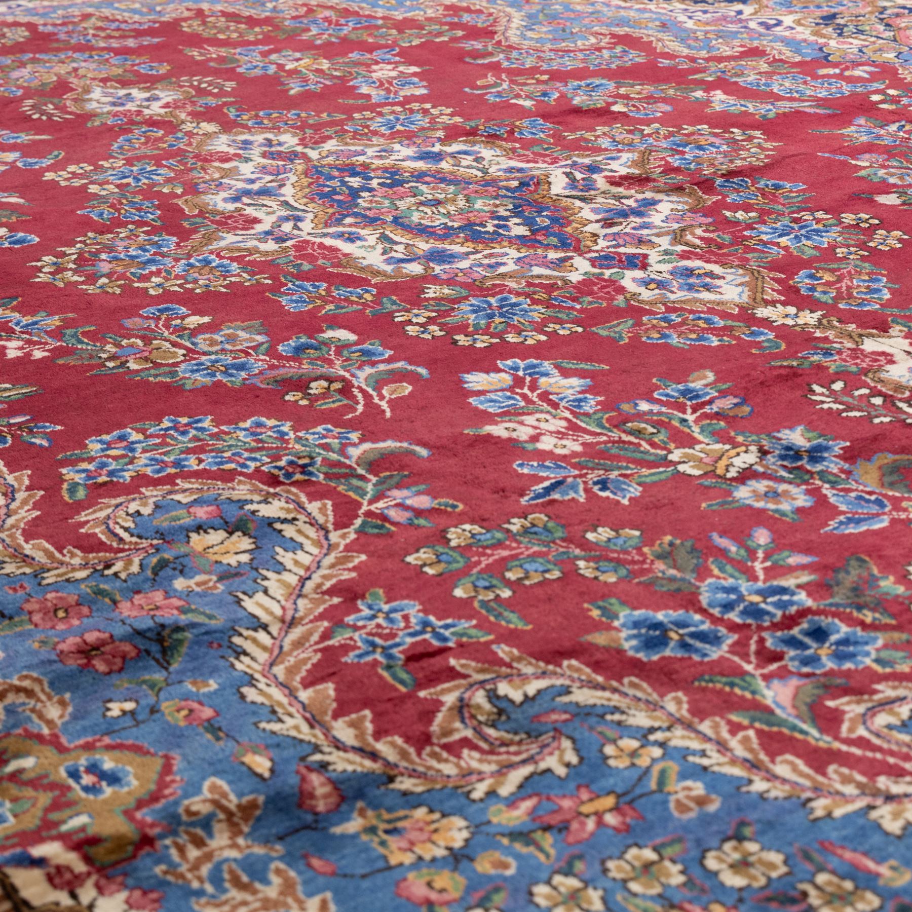 Large Central Persian Kirman hand-knotted carpet, raspberry red field, with central shaped medallion surrounded by an intricate arrangement of scrolling vines and floral sprays, within conforming spandrels and a pale blue border decorated with floral motifs, enclosed by guard stripes, with cotton fringe ends 