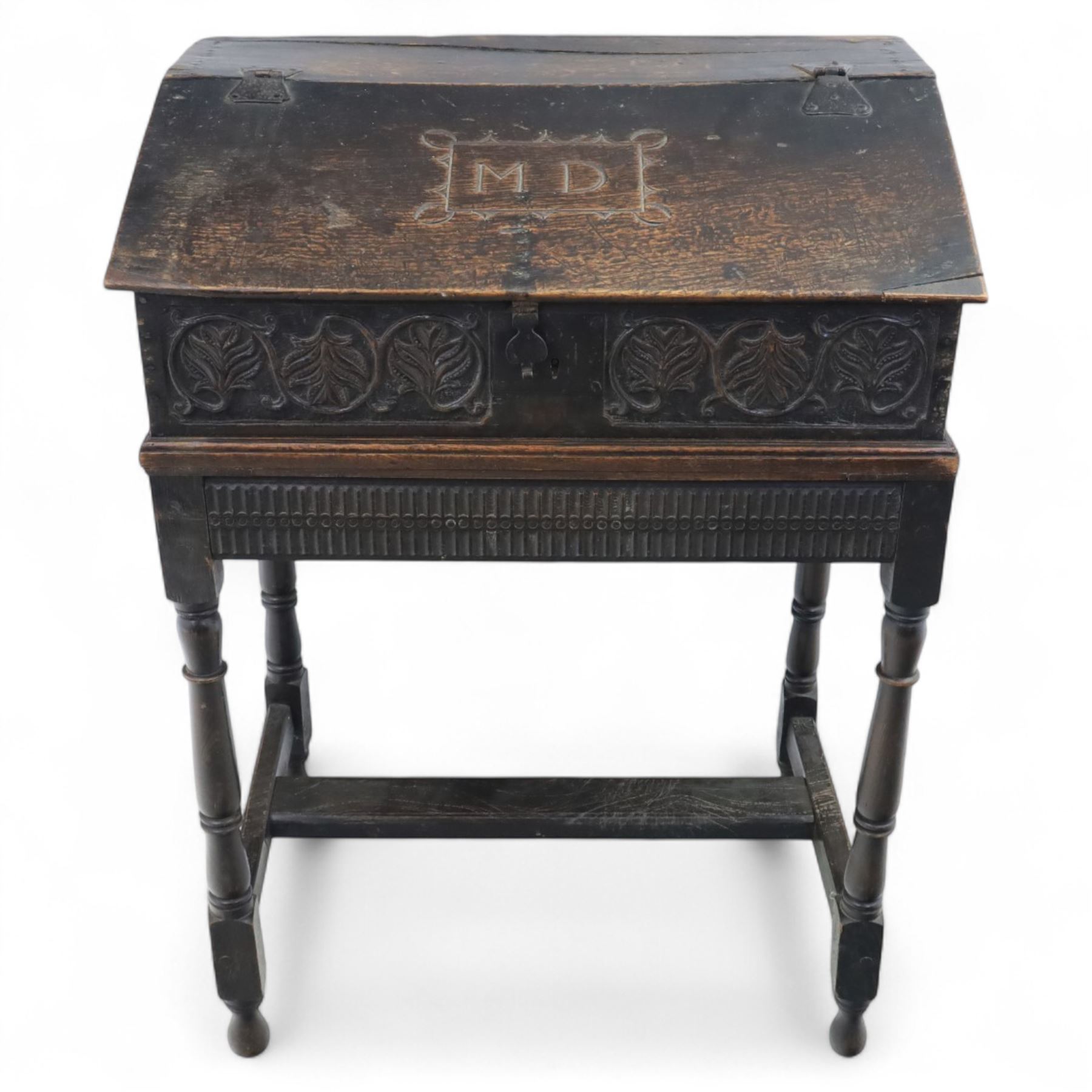 18th century carved oak Bible box on stand, the sloping lid with incised initials ‘MD’ and iron strap hinges, the front carved with stylised leaf and scroll panels enclosing central iron lockplate, on stand with gadrooned frieze, turned legs and plain stretchers