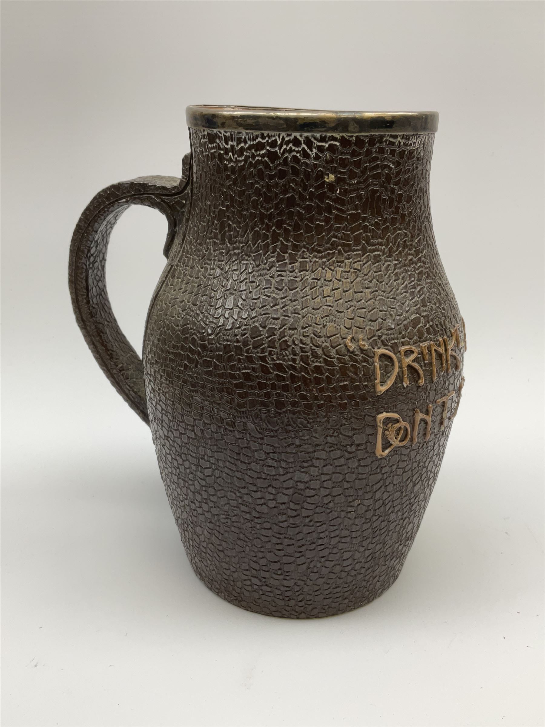 A Doulton Lambeth stoneware jug, with silver mounted collar, hallmarks rubbed, maker's mark probably Cornelius Desormeaux Saunders & James Francis Hollings (Frank) Shepherd, the leather effect body titled 'Drinke Faire Don't Sware', with impressed marks beneath including 'Manufactured for Sidney W Allen 39 White Rock Hastings', H18.5cm. 
