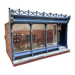 Good quality late 20th century dolls house by M. James dated 1995 in the form of a Beverley East Yorkshire Antique Shop called 'North Bar Antiques'; single-storey single-roomed shop with textured brick walls and flat roof, fully glazed hinged front with recessed hinged customer door and central window to the rear; comprehensively furnished with chests of drawers, pedestal desk, breakfast table, lowboy, two longcase clocks, delft rack with ceramics, occasional tables, pictures, Regency style mirror and collectibles; decorated walls with coving and picture rail and simulated floorboarding; wired for electric lighting; maker's plaque W61cm H42.5cm D41.5cm