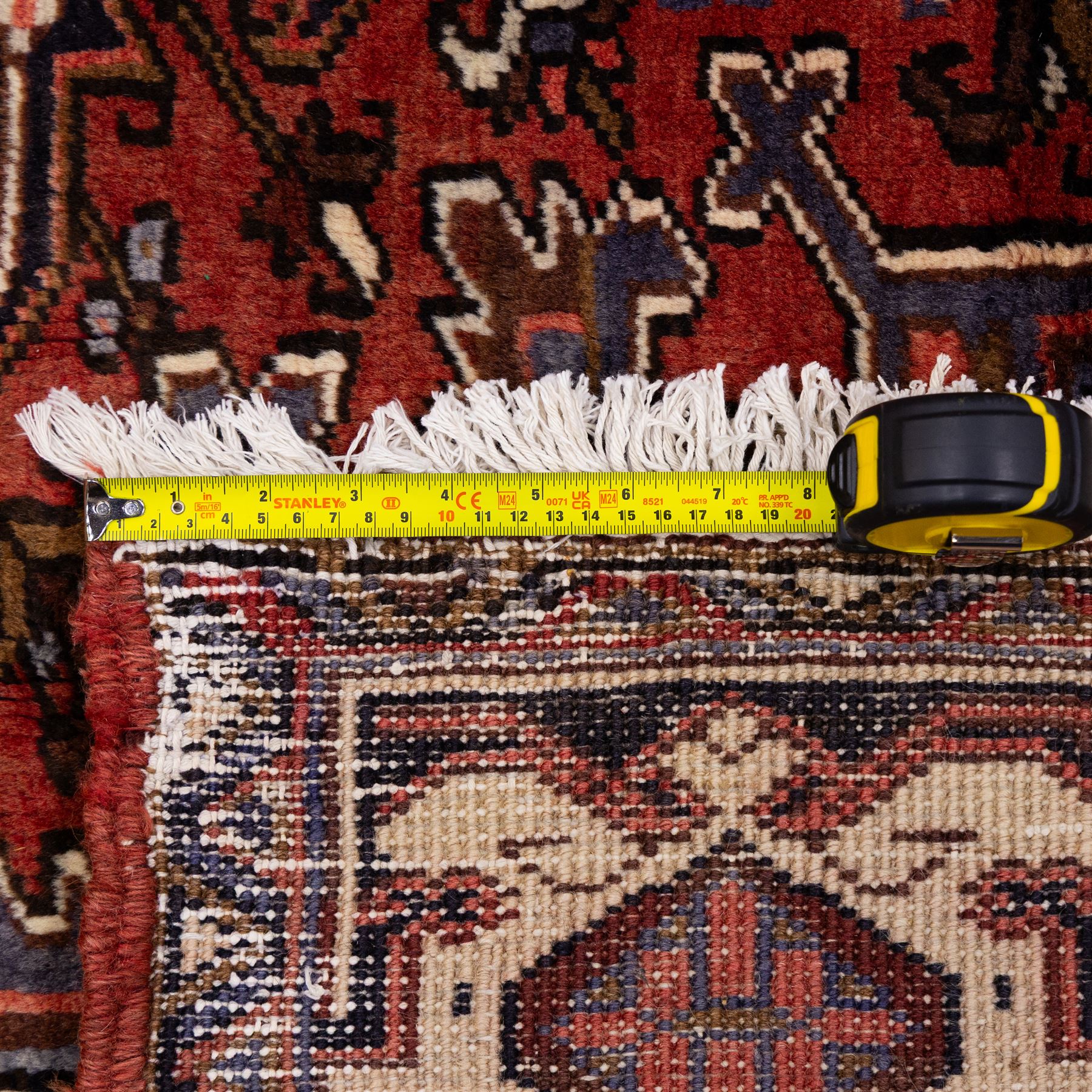 North West Persian Heriz hand-knotted carpet, madder red field with central stepped geometric medallion with radiating angular motifs and serrated outlines, and stylized floral and tribal devices framed by a broad border of repeating angular palmettes and rosettes flanked by multiple guard stripes, with cotton fringe ends 