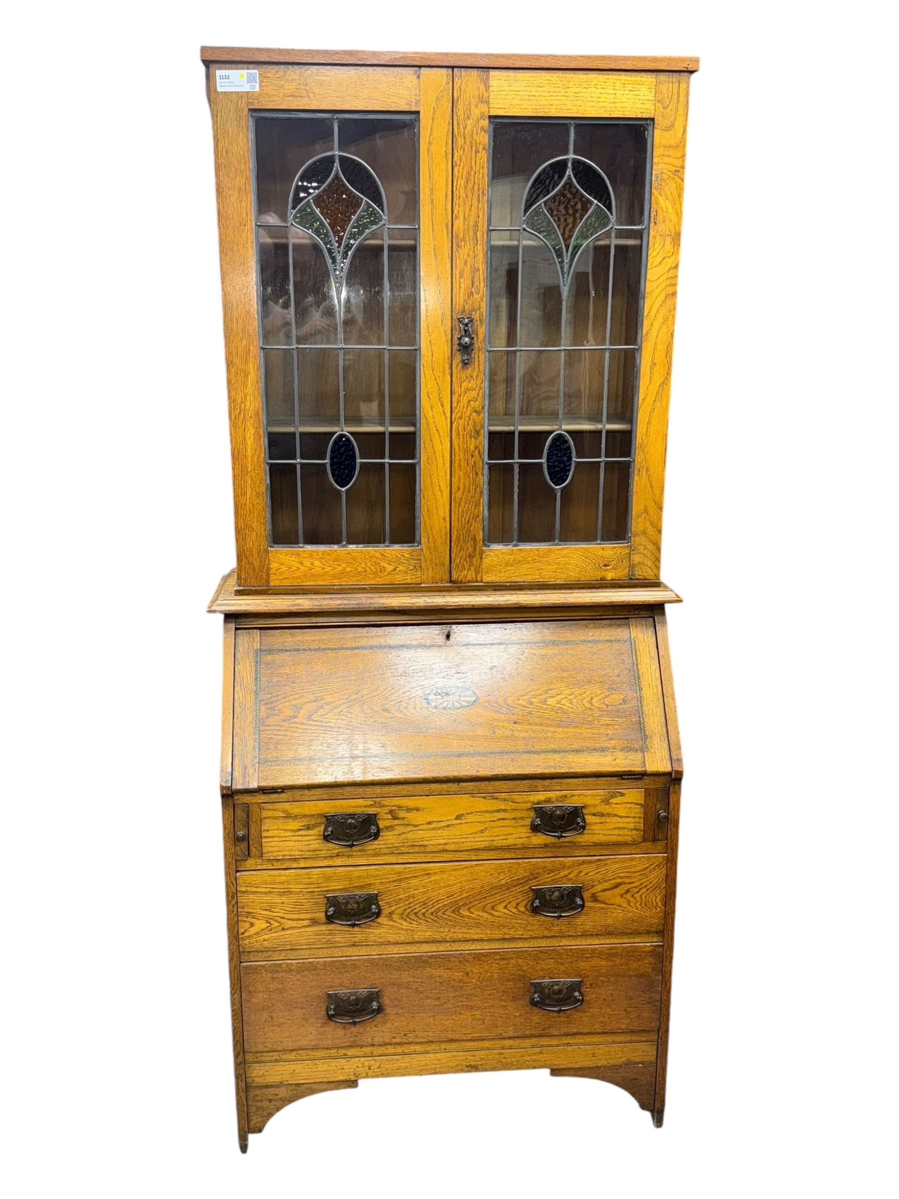 Early 20th century Arts and Crafts oak bureau bookcase, twin leaded and glazed stained glass doors enclosing two shelves, over bureau section with fall-front inlaid with sunburst motif, fitted interior of pigeonholes and shelves, on bracket feet