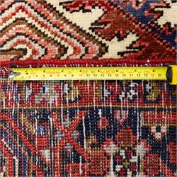 North West Persian Heriz hand-knotted carpet, madder red field, with a large angular stepped central medallion with serrated outlines, flanked by bold stylized palmettes, hooked leaves and geometric devices, spandrels similarly decorated within a wide geometric main border featuring angular floral motifs, enclosed by multiple guard borders in madder red, indigo blue, ivory, olive green and soft ochre, with cotton fringe ends 