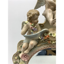  A late 19th century Sitzendorf porcelain mirror, the mirror plate of circular form set within a shaped surround encrusted with flowers and foliage, surmounted by three figures of cherubs and raised upon four scroll feet, with blue cross hatch mark beneath, H57cm.   