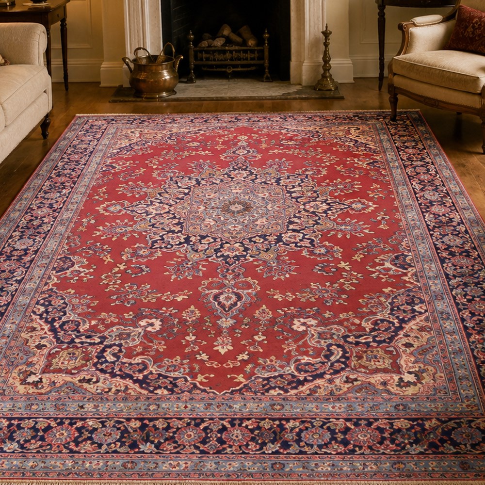 North East Persian Meshed hand-knotted rug, madder red field with central multi-lobbed medallion in shades of indigo, ivory, and sofa pastel tones surrounded by scrolling floral vines, palmettes, stylized blossoms, floral sprays, and cartouches, framed by Indigo spandrels, enclosed within a broad dark blue main border, flanked by multiple narrow guard stripes, with fringe ends 