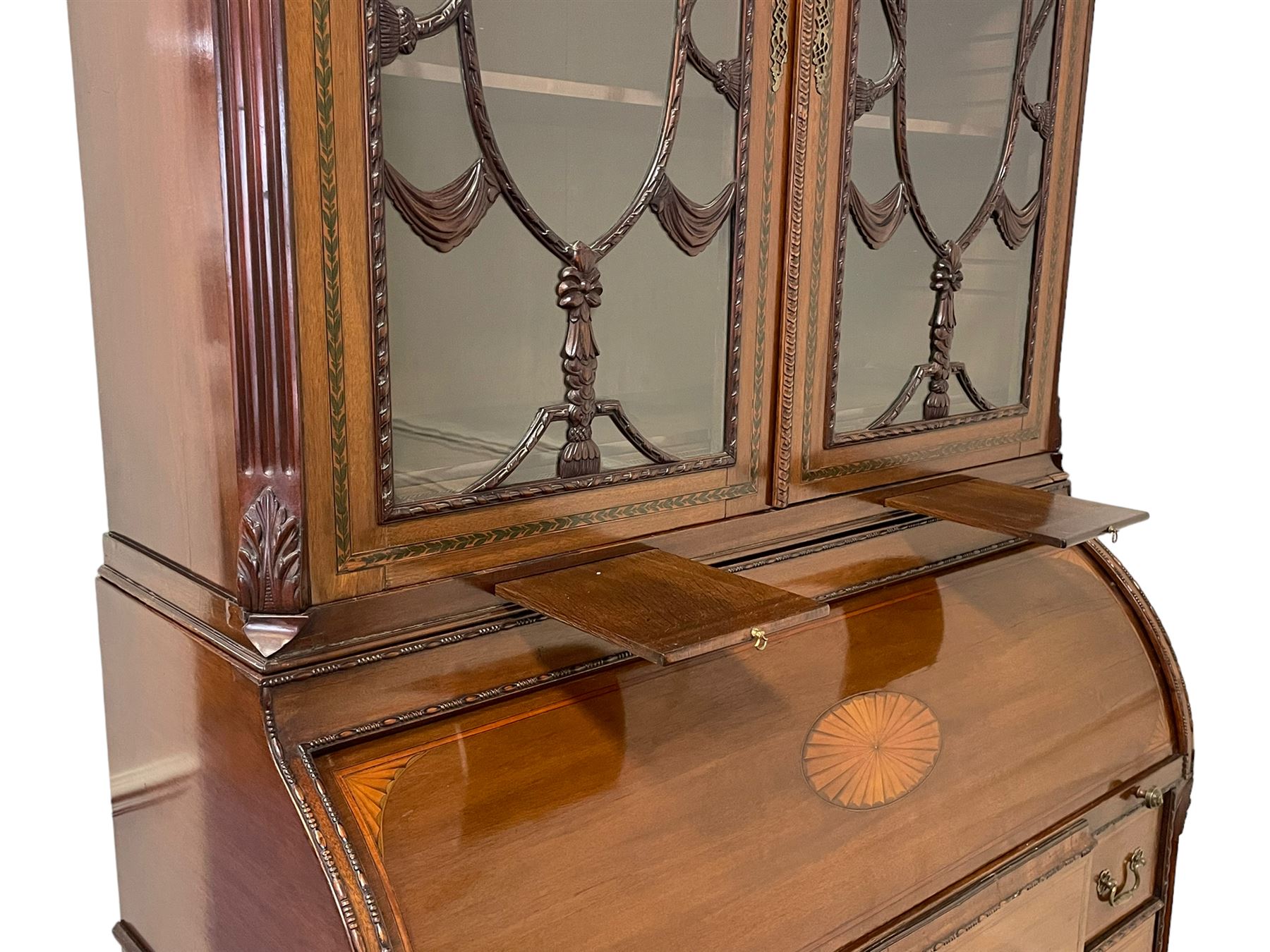 Large and impressive late 19th century Irish mahogany secretaire bookcase, moulded sloped arch dentil pediment decorated with central carved mask surrounded by linen festoons and trailing bellflowers, canted and fluted uprights with carved acanthus leaf upper and lower brackets, enclosed by two astragal glazed doors in the form of trophies with twist ribbon carved mouldings, inlaid with fanned spandrels and trailing leaf painted satinwood band, cylinder roll top inlaid with central fan motif and fanned spandrels, pull-out action revealing stepped fitted interior, central tambour roll cupboard flanked by two Composite pilasters and a combination of drawers and pigeon holes, fitted with three break-front drawers, matched canted and fluted corners with acanthus leaf carvings, on square tapering and fluted spade feet