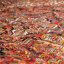 North West Persian Heriz hand-knotted carpet, madder red ground, with a large central geometric medallion with pendants, surrounded by angular foliate and floral motifs incorporating stylised rosettes, palmettes and serrated vine work, enclosed by ivory spandrels and a dark navy border with repeating geometric flowerheads and angular meandering vines, flanked by narrow patterned guard stripes, with cotton fringes