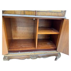 Early 20th century walnut veneered cocktail cabinet, shaped and carved foliate pediment above a pair of arched panelled doors with figured veneers and ornate cast escutcheons, interior is mirrored on the base and back, slide out mirrored shelf, flanked by foliate carved uprights, over a conforming lower cabinet, all raised on shaped apron and scroll carved cabriole feet