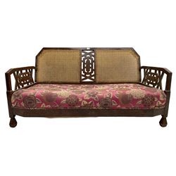 Early 20th century two seat sofa, cane back, the central panel and arm supports pierced with a geometric design, the sprung seat upholstered in fuchsia floral patterned fabric