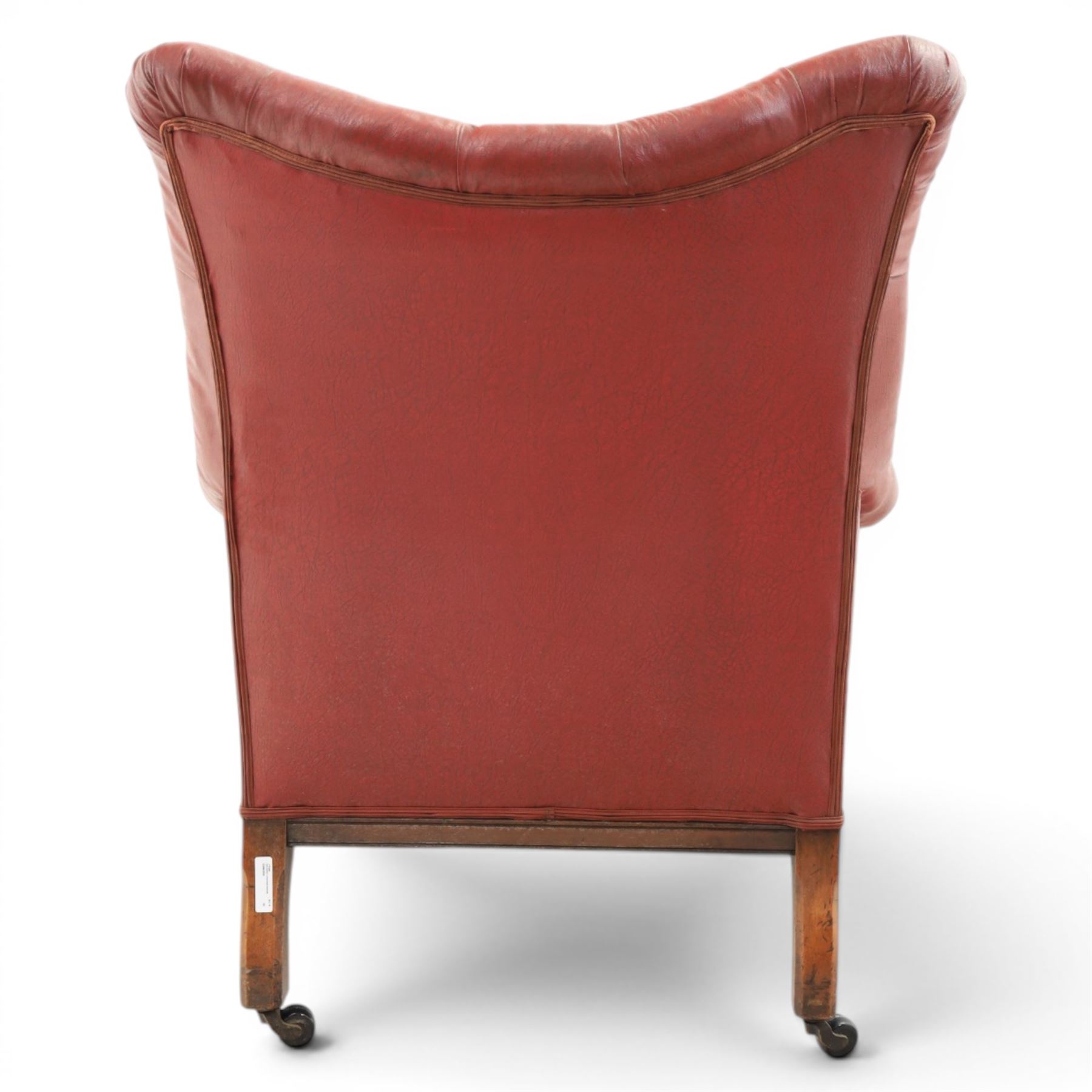 Early 20th century style oxblood red leather Chesterfield armchair, deep buttoned shaped back over padded scroll arms and loose seat cushion, raised on turned mahogany front supports with brass caps and castors