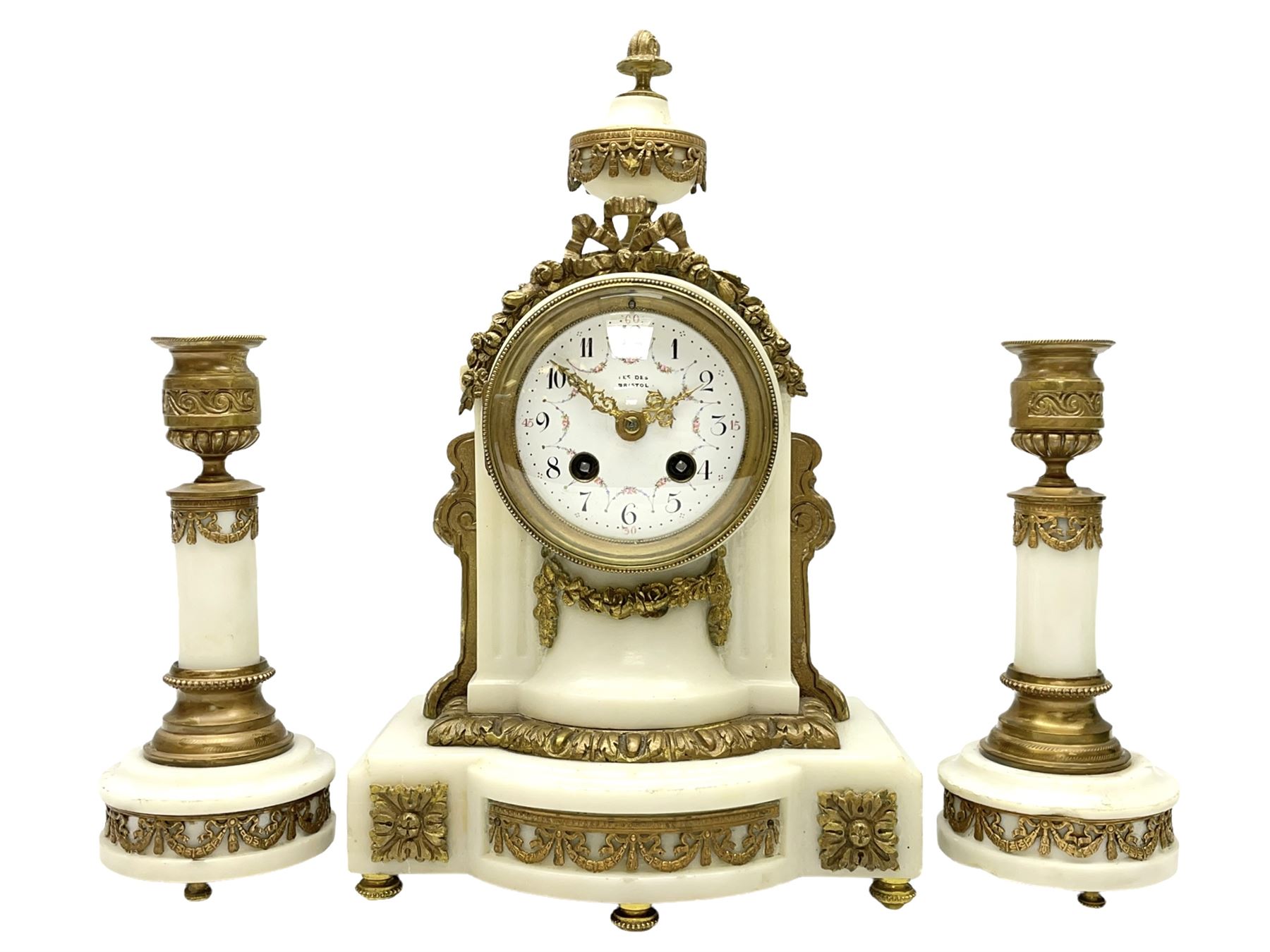 An early 20th century white marble mantle clock in a break front case with an arched top and finial, gilded scroll work to the sides and pierced foliate panels to the front, plinth raised on five bun feet, enamel dial with garland decoration, Arabic numerals, minute markers and Louis XV gilt hands, within a convex glazed bezel, eight-day Parisian striking movement striking the hours and half hours on a bell, with two conforming single light candlesticks. With Pendulum.

