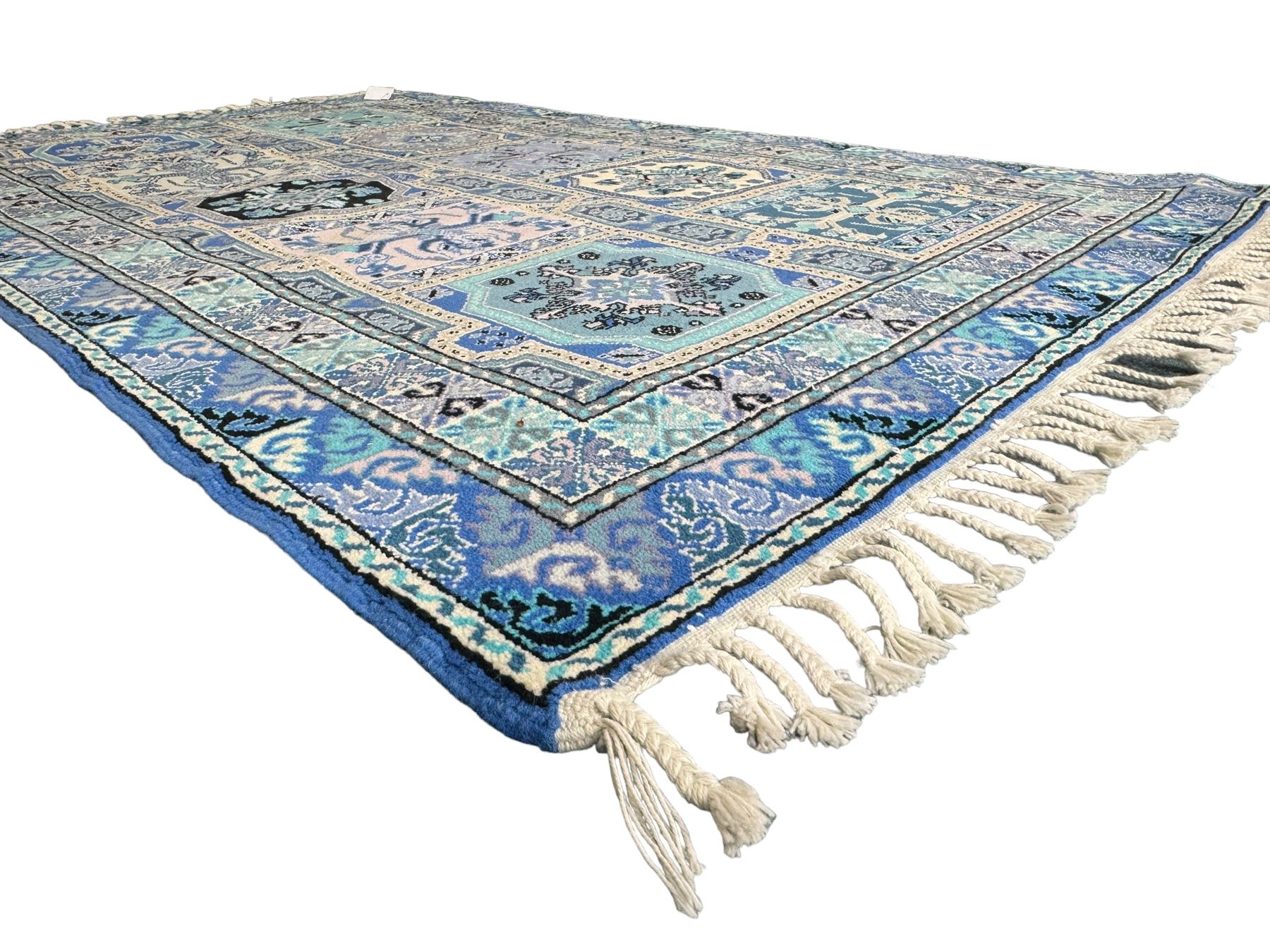 Morrocan single weft blue ground rug, the field segmented into a grid of ten rectangular panels, bordered by a series of guard bands with repeating floral and geometric patterns with interlocking vines and rosettes, the edges finished with a fringe of twisted tassles
