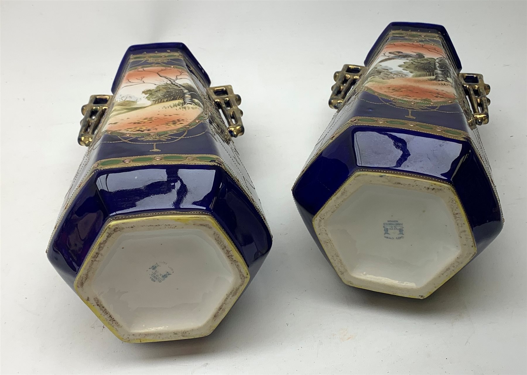 A pair of Japanese Nippon Kinjo vases, of tapering hexagonal form with twin stylised handles, the cobalt blue ground with oval hand painted panels to front and verso depicting landscapes, further detailed with smaller floral bands and panels and heightened with gilt, each with mark beneath, H30.5cm.  