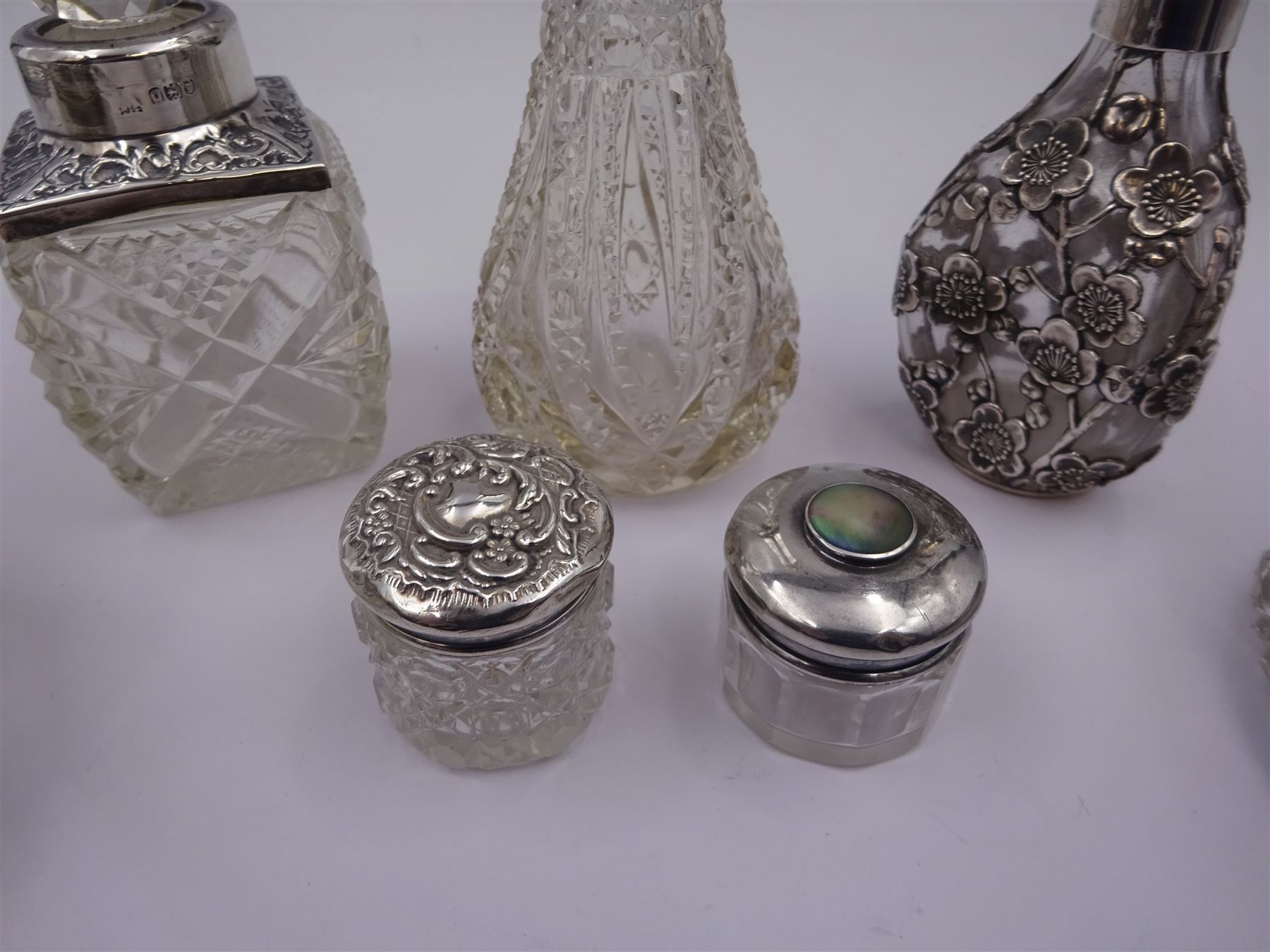 Two silver mounted dressing table jars, one in the form of a heart, both with embossed silver covers and cut glass bodies, hallmarked, together with two smaller examples, one set with mother of pearl, three silver mounted glass scent bottles, including an example with prunus blossom decoration, stamped Sterling, tallest bottle H17cm