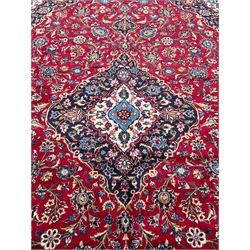 Central Persian Kashan crimson ground rug, the floral ivory and indigo medallion surrounded by palmette motifs with scrolling foliate branches, the guarded border decorated with repeating stylised flower heads