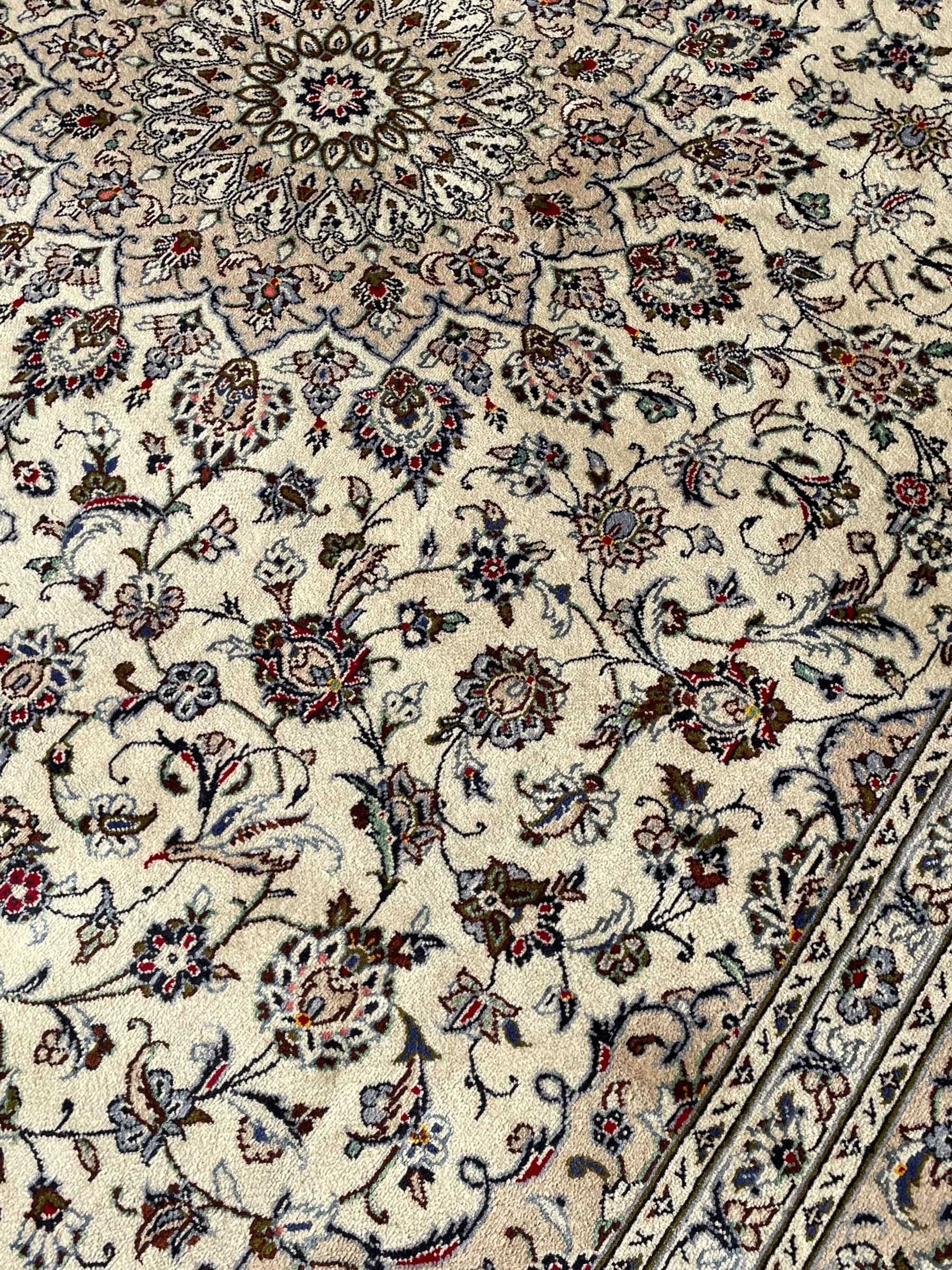 Persian Kashan ivory ground carpet, central rosette medallion with projecting palmettes, densely decorated field depicting trailing leafy branches and stylised plant motifs, the border with similar scrolling pattern, within floral pattern guard stripes 