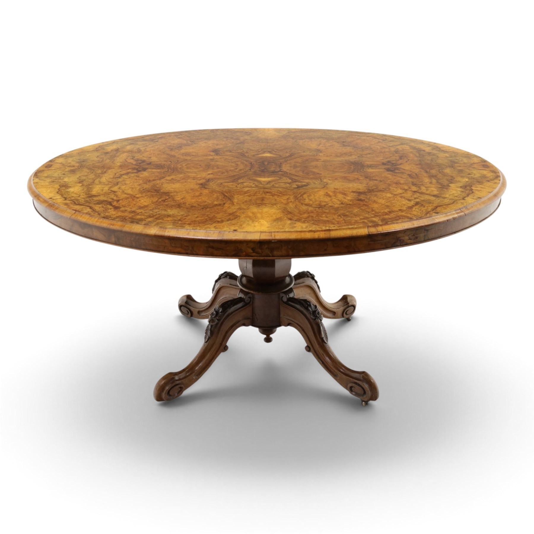 Victorian figured and burr walnut loo table, oval book-matched veneer top with moulded edge, over a turned octagonal pedestal terminating to four splayed supports with flowerhead carved knees and scroll feet with castors