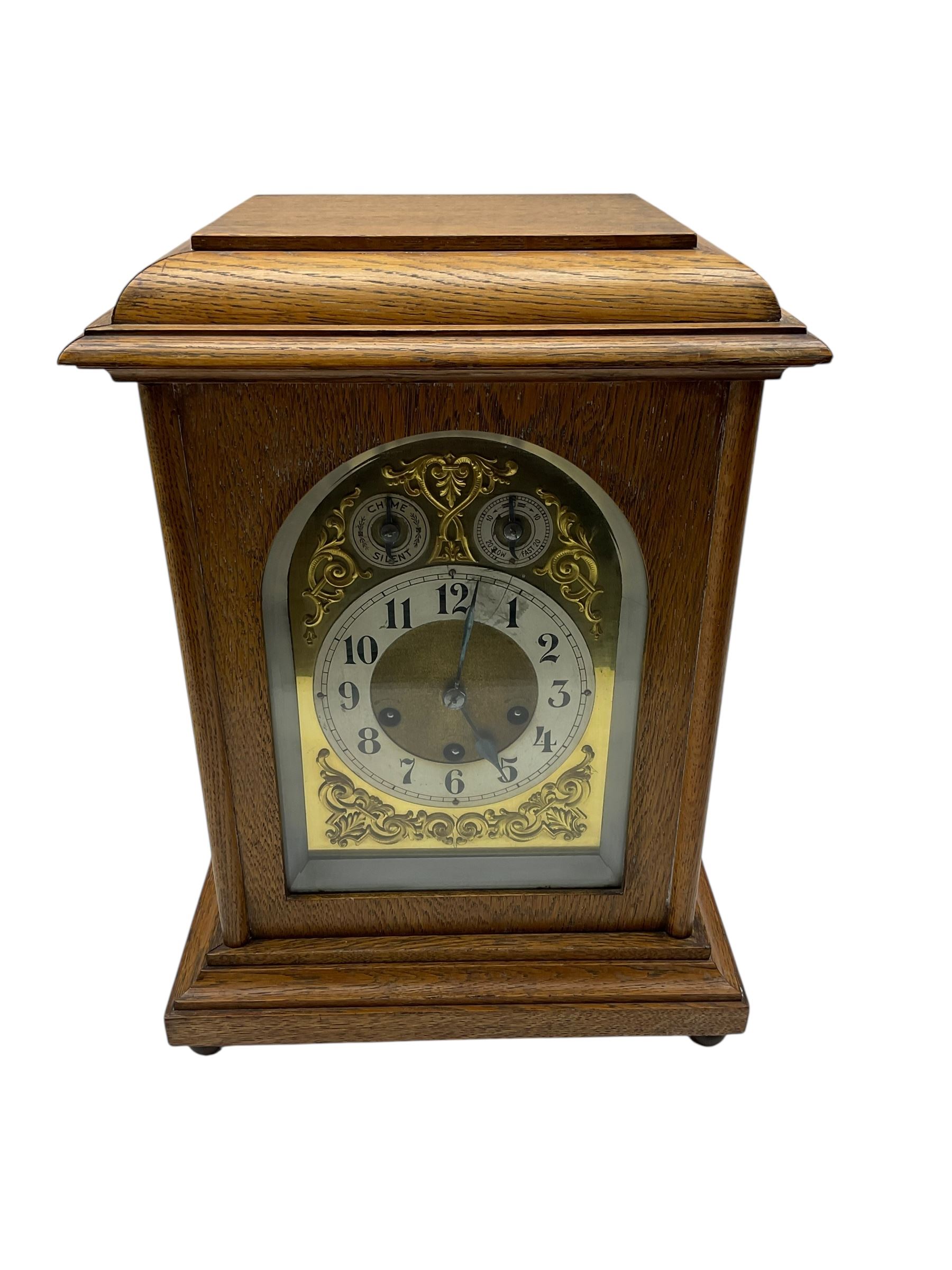 Jungans-German Edwardian 8-day oak cased mantle clock, with a stepped flat top and glazed door on a shallow plinth with bun feet, brass dial with cast spandrels, silvered chapter with Arabic numerals, chime/silent and pendulum regulation dials, three train spring driven movement sounding the quarters on gong rods and the hours on a coiled gong. With pendulum.