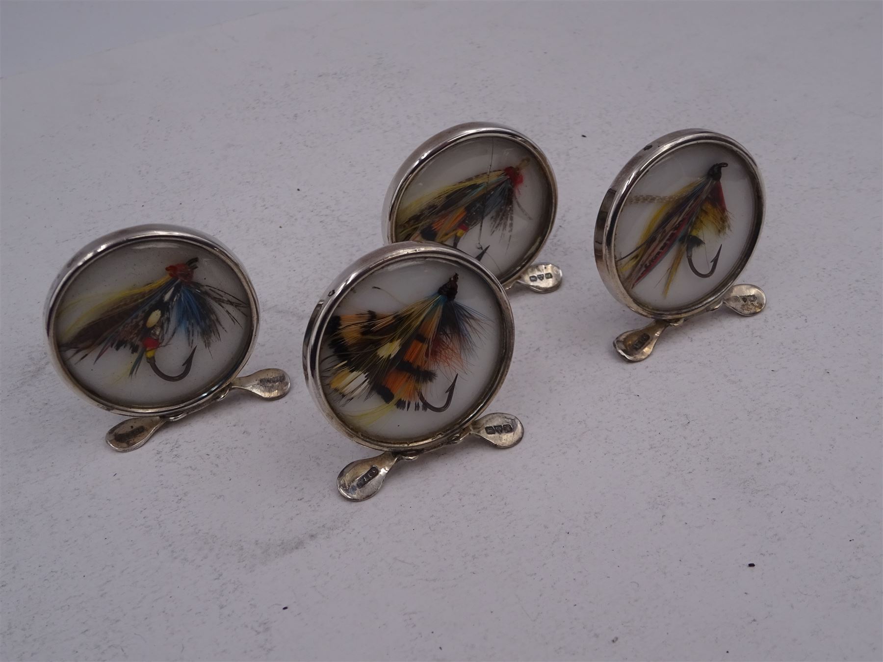 Set of four early 20th century silver mounted place card holders, each of circular form and set with a coloured fly fishing hook behind a glazed panel, upon three pad feet, hallmarked Grey & Co, Chester 1912, H4.5cm, contained within red fitted case, with purple silk and velvet interior