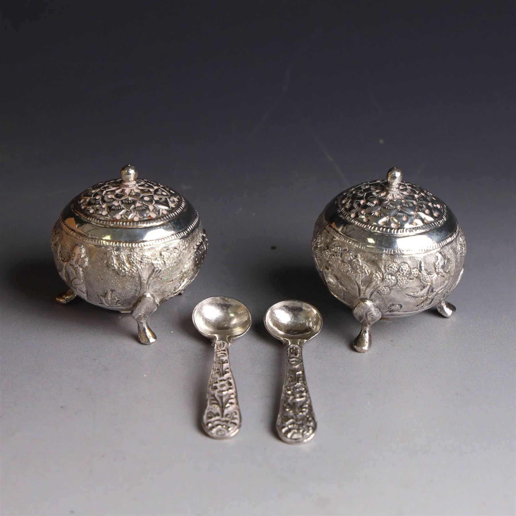 Pair of Middle Eastern silver cruets, of spherical form with pierced tops, embossed with figures, elephants and village scenes, unmarked, together with a pair of silver condiment spoons embossed with floral decoration 