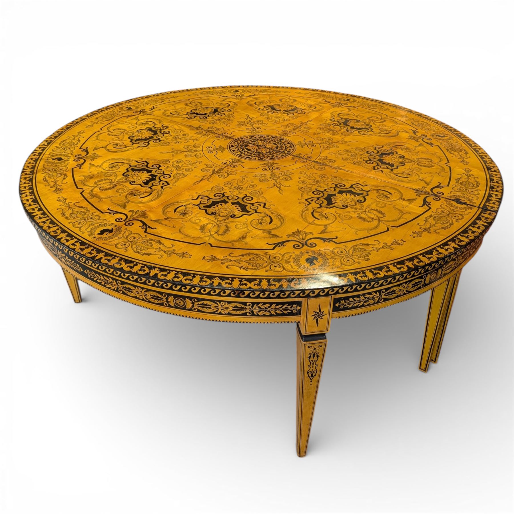 20th-Century Louis XVI-Style satinwood and penwork circular table or pair of console tables, the circular quarter-veneered top decorated in penwork with a central classical mask head motif within scrolling foliate and anthemion ornament on a contrasting black ground, above a conforming frieze decorated with Greek key-type scrolls. Raised on square tapering legs, each with an upper block panel decorated with compass-star motifs.