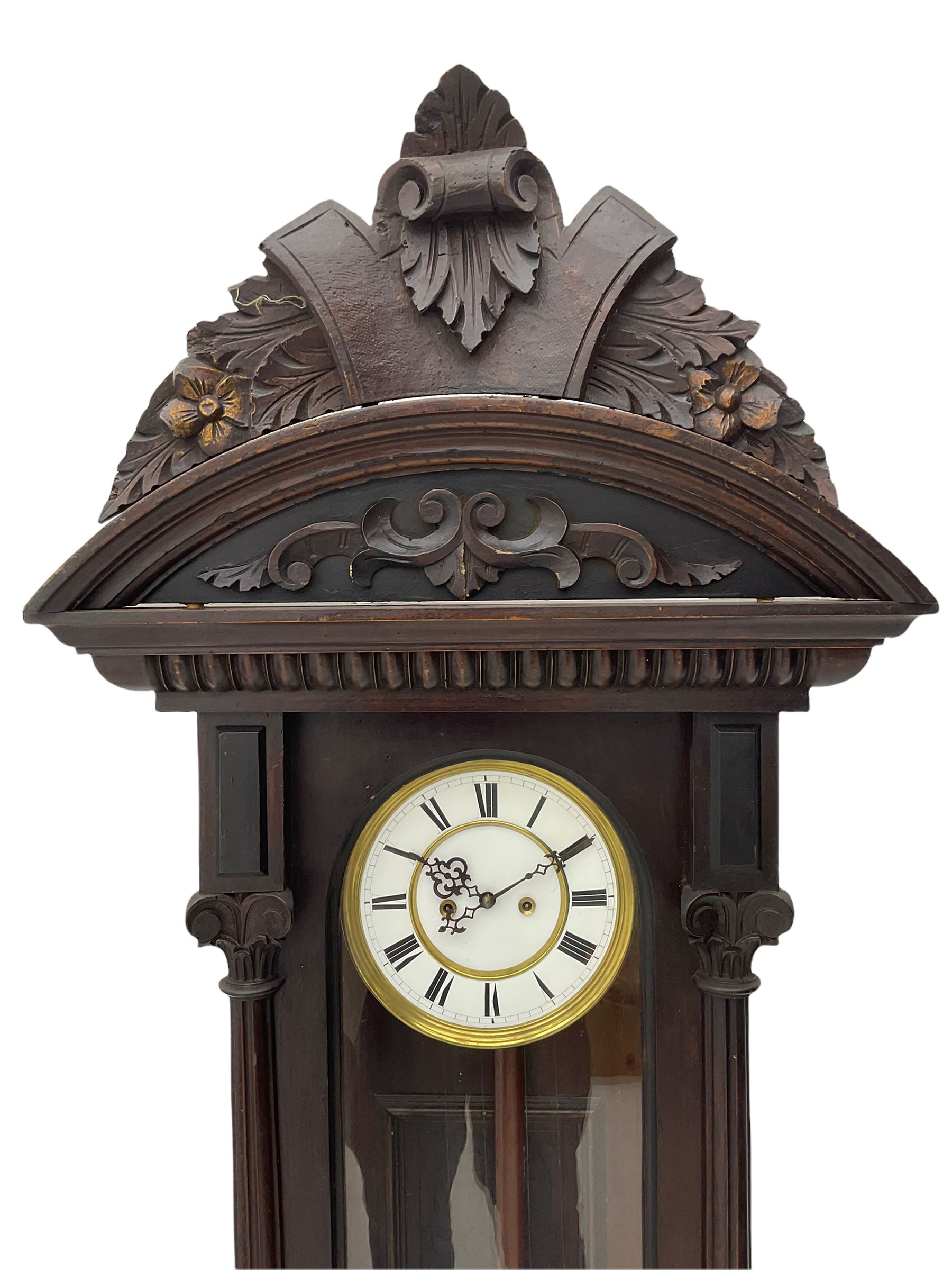 A late 19th century German Vienna style weight driven wall clock c1880, with a predominant arched pediment and applied carving, case with glass side panels (one missing) and a glazed full-length arched door flanked by reeded columns with decorative capitals, 7' two-piece white enamel dial with roman numerals, minute markers and pierced steel gothic hands, pendulum with a wooden rod, pressed brass bob and beat plate, eight-day weight driven movement striking the hours and half hours on a coiled gong. With brass cased weights.



