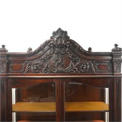 Large and impressive 19th century heavily carved mahogany display cabinet, the ornate pediment carved with a central scalloped and foliate rocaille motif with extending acanthus leaves and egg-and-dart moulding flanked by acanthi finials, over a central cartouche with carved bow-and-arrow motif and trailing scrollwork, the bookcase enclosed by two glazed doors with bevelled glass sides, the base with a gadrooned upper edge over a central rocaille ornament with scrolling acanthus leaves, the panelled frieze with further foliate motifs, raised on large carved snarling lion mask supports with floral garlands and claw feet, united by a concave rectangular platform base with gadrooning egg-and-dart moulding