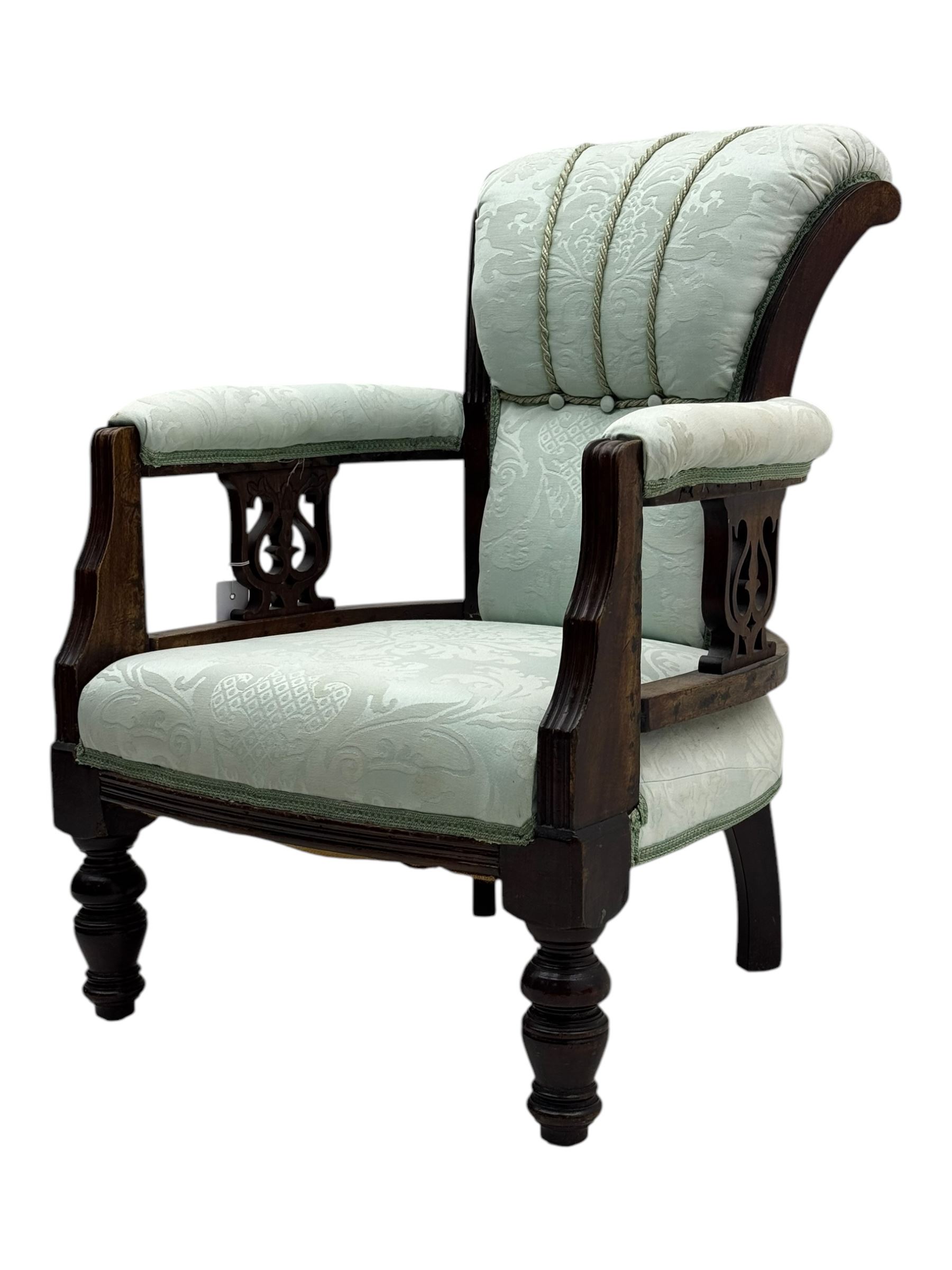 Late Victorian carved mahogany open armchair, the shaped back with fluted upholstered panel and padded arms above pierced scroll carved side supports, over a broad upholstered seat, on turned front supports
