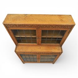 Early 20th-century Arts and Crafts glazed oak bookcase, upper section fitted with a pair of leaded glazed doors enclosing a fixed shelf above a projecting frieze drawer interior with baize-lined writing surface flanked by two compartments, carved with interlaced decoration, lower section fitted with a further pair of leaded glazed doors enclosing a single shelf, raised on short stile feet 