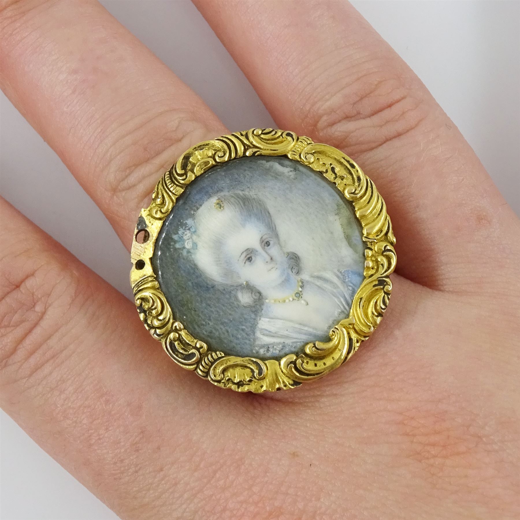 19th century gold portrait ring, set with a circular miniature of a young woman in 18th century dress on ivory, under a glazed panel, within a gold scroll frame and pierced gallery. This lot has been registered for sale under Section 10 of the APHA Ivory Act