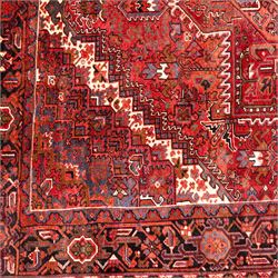 North West Persian Heriz hand-knotted carpet, madder red field, with a large central angular star medallion with stepped outlines on ivory ground, surrounded by geometric floral devices and stylized rosettes, with corresponding corner spandrels, enclosed by a bold geometric main border with multiple guard stripes, with cotton fringe ends 