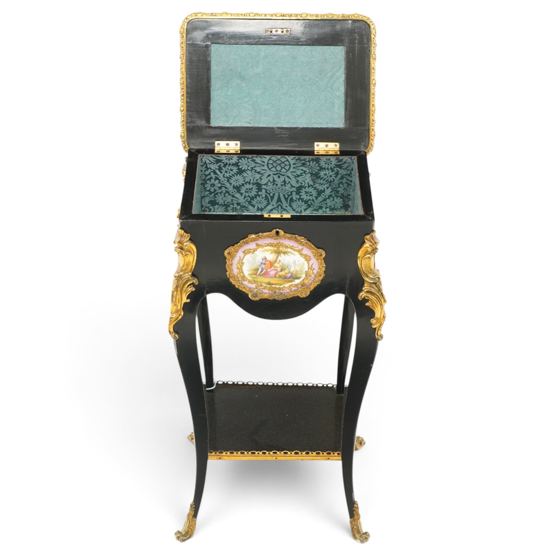 Early 20th century French ebonised sewing or work table, rectangular hinged top with foliate moulded gilt metal edge, enclosing upholstered interior, over a central Sevres style porcelain plaque depicting a courting scene, raised on cabriole supports with gilt metal carotuche and acanthus knee mounts, united by undertier with brass gallery terminating to gilt metal scroll feet