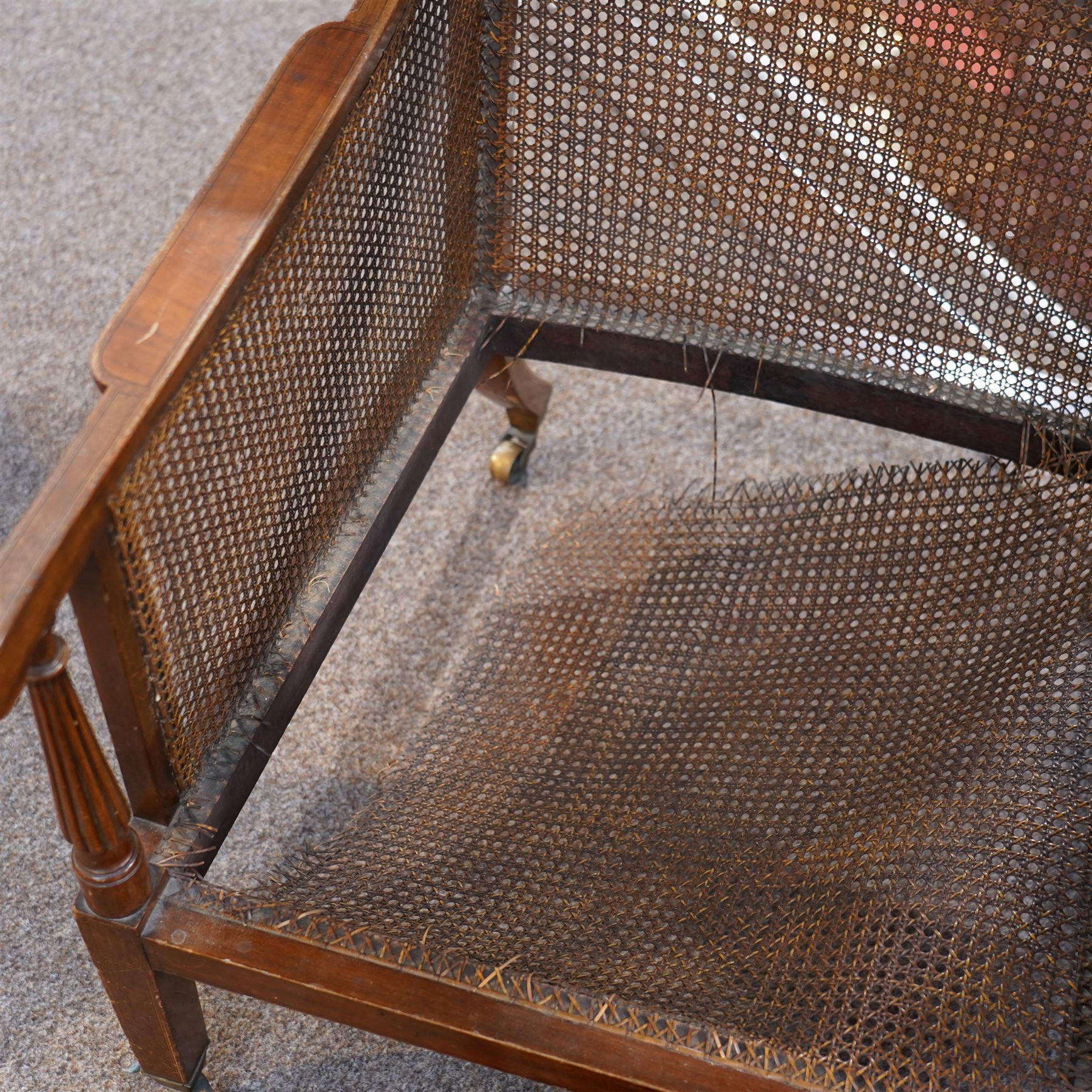 Pair of early 20th-century mahogany Bergère armchairs in the manner of Gillows, the back and seat with woven cane panels with loose red velvet cushions, the frame with swept arms with tapered and reeded uprights, raised on tapered square-section front legs terminating in brass cup castors 
