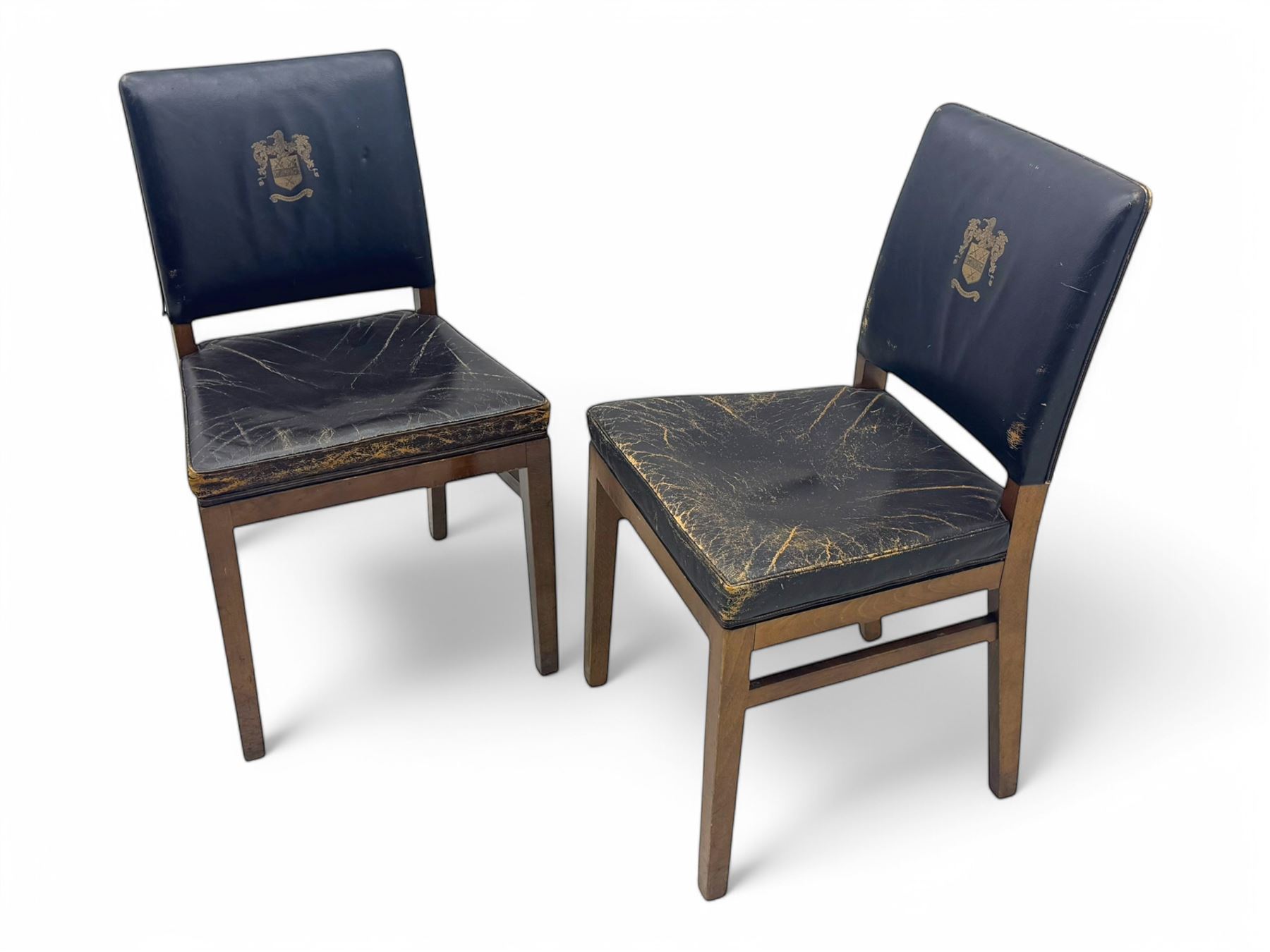 Pair of mid-20th century hardwood chairs associated with the Company of Cutlers in Hallamshire, Sheffield, each featuring a squared frame and black vinyl upholstery, the back emblazoned with the guild’s gilt coat of arms and motto “Pour Y Parvenir a Bonne Foi” (To Succeed through Honest Endeavour)